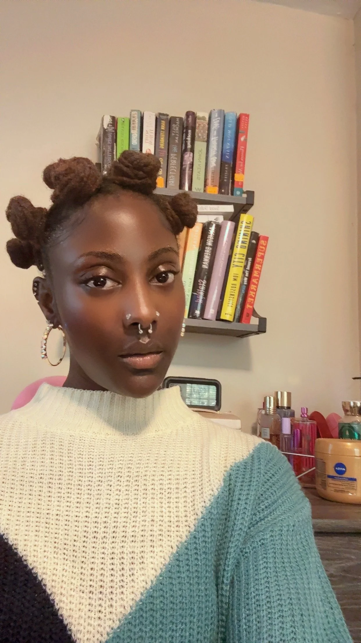 A young woman with styled brown bantu knots, wearing hoop earrings and a striped sweater, taking a selfie in a room with bookshelves, perfume bottles, and a small desk in the background.