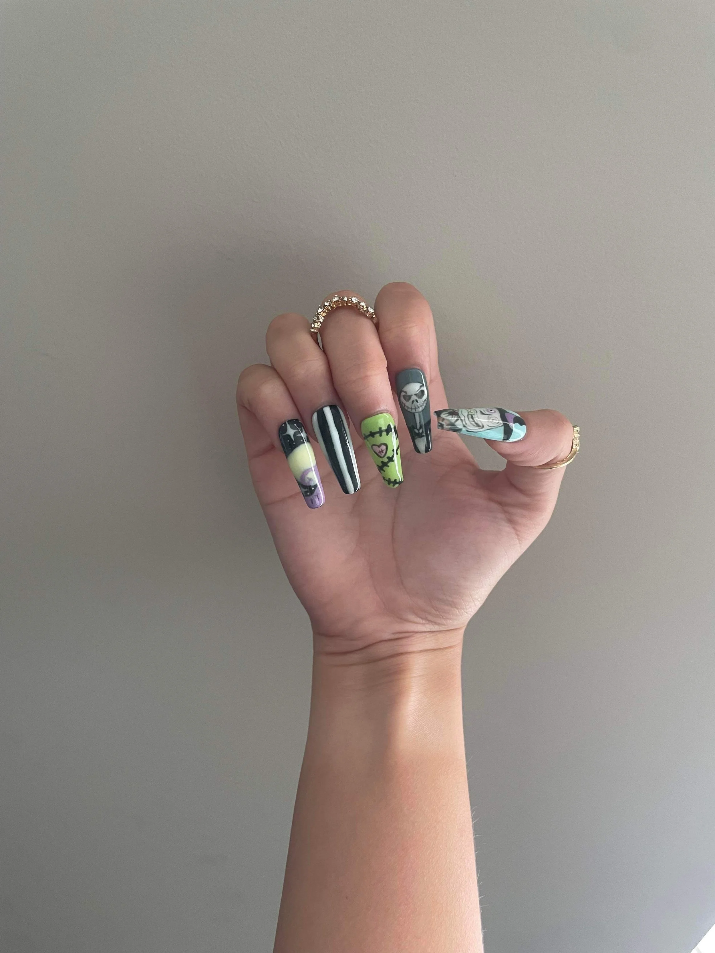 A hand with decorated Halloween-themed nails, showcasing a silver ring on the middle finger and a gold ring on the ring finger, set against a plain gray background.