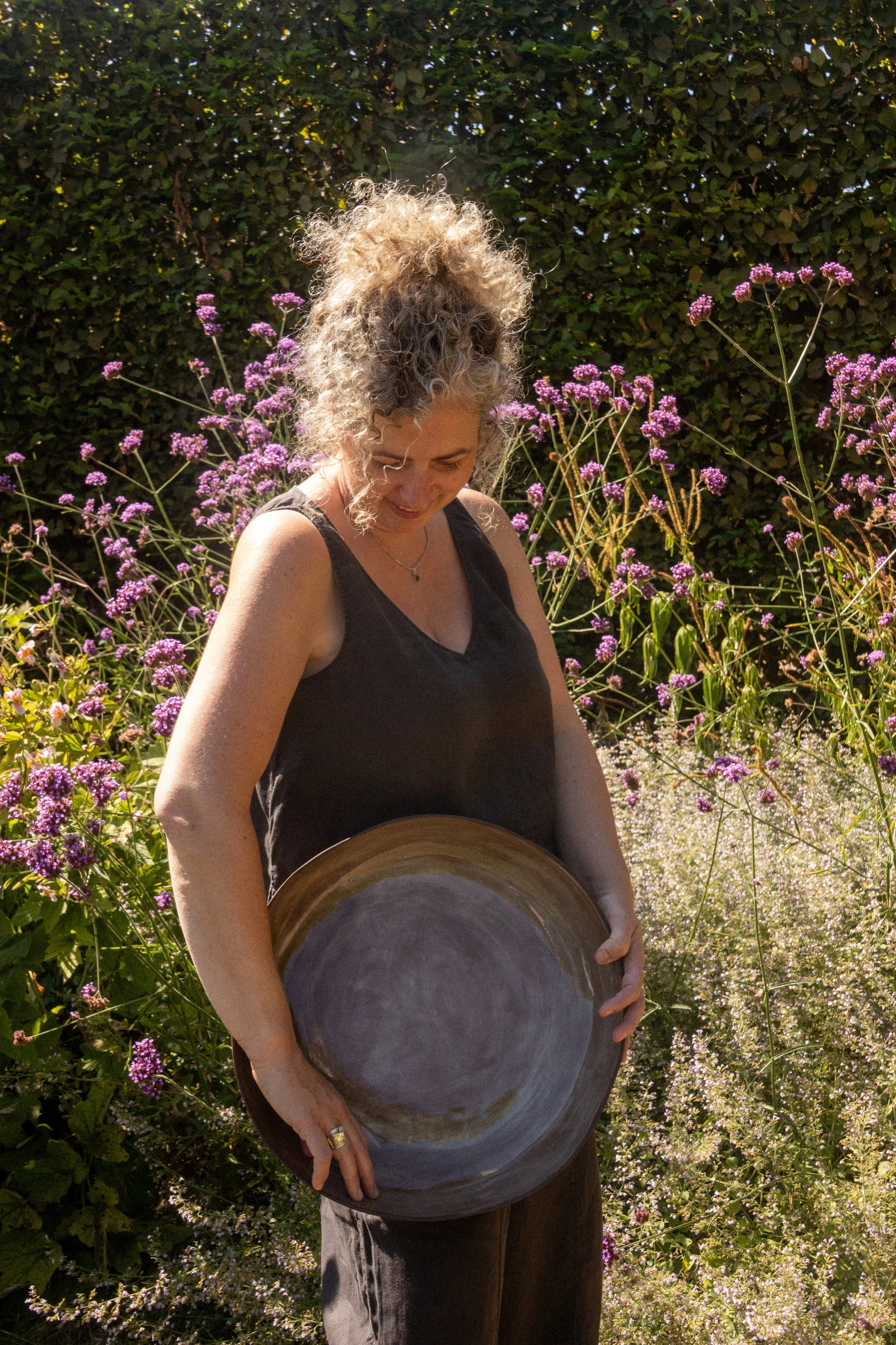 Een vrouw met gekruld blond haar in een tuin, omgeven door paarse bloemen, die een grote keramische pot vasthoudt.