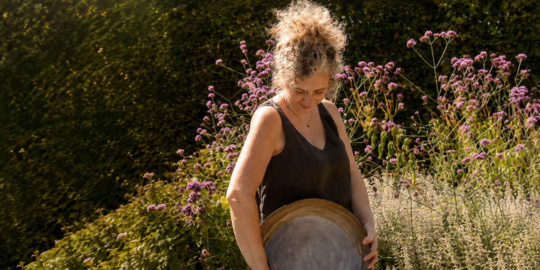 Een vrouw met krullend blond haar die een grote, lege metalen schaal vasthoudt in een tuin met bloeiende roze en witte bloemen.