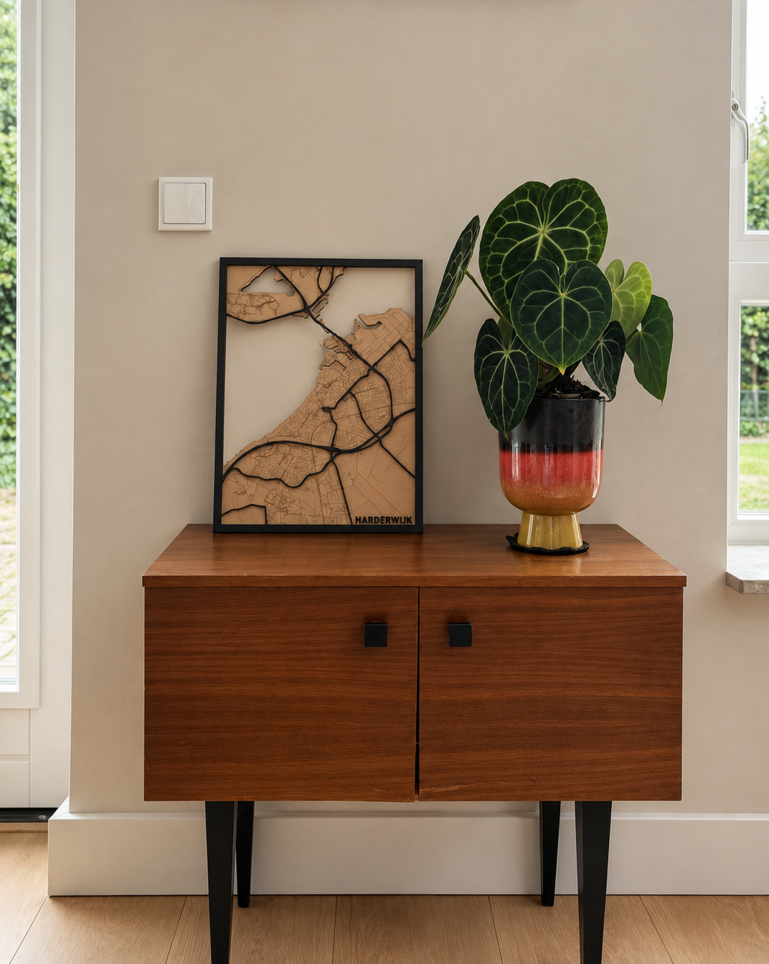Een houten kast met zwarte poten, een wereldkaart en een potplant met grote groene bladeren op een beige muur.
