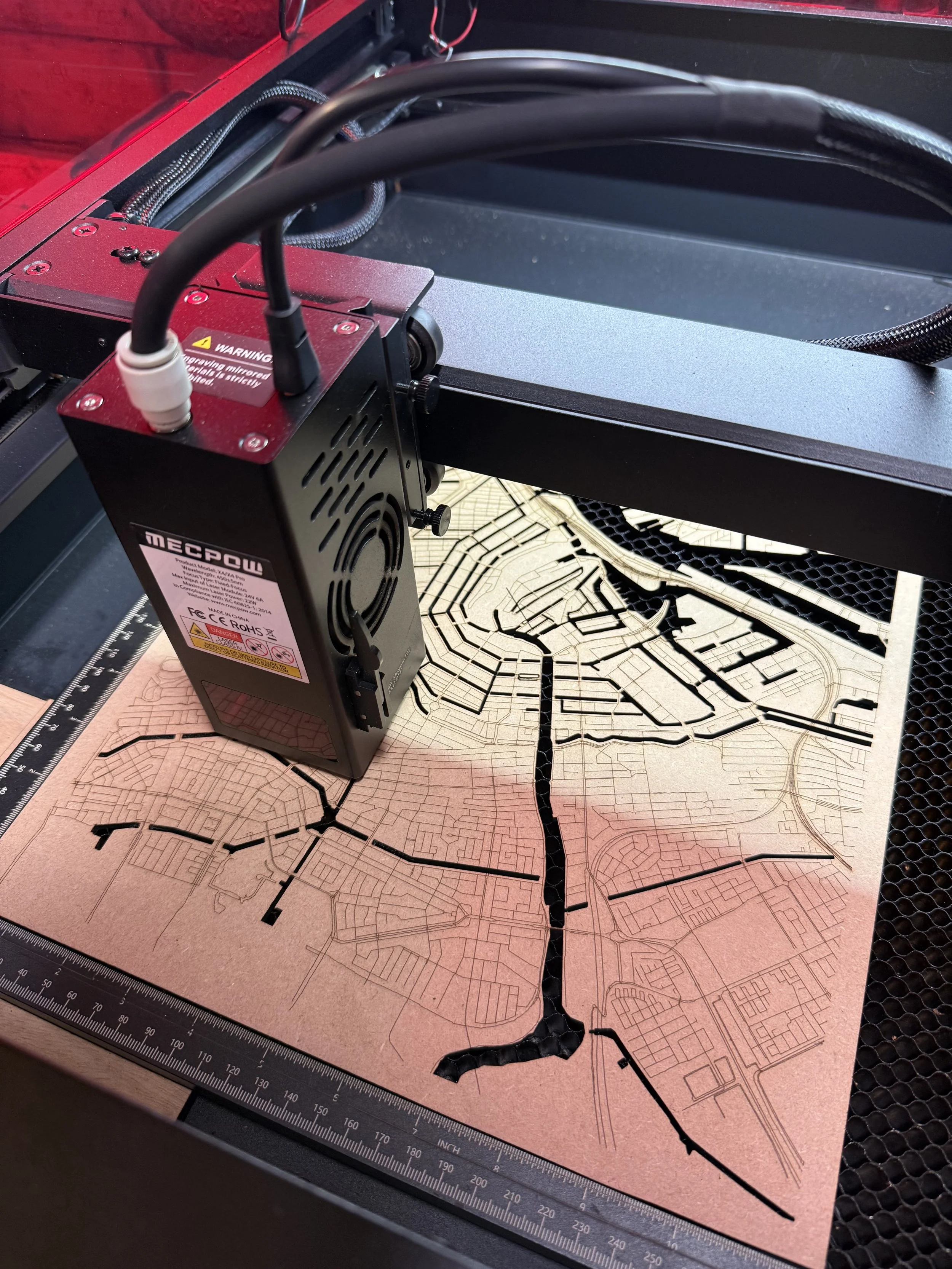 Een 3D-printer die een plat houten kaartje met een stratenplan van een stad print, onder een metalen meetlint. De printer is zwart met rode accenten en bevindt zich in een werkruimte met een ruit.