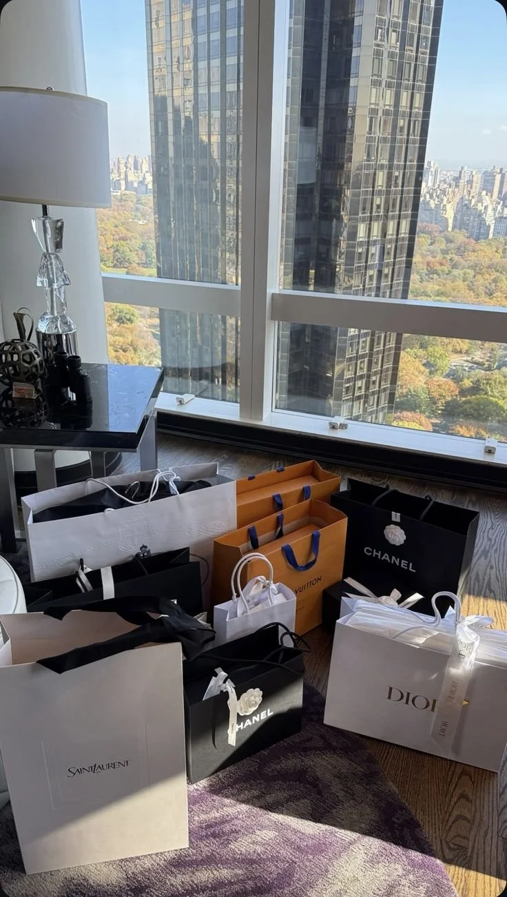 Multiple luxury shopping bags from brands like Chanel, Louis Vuitton, Saint Laurent, and Dior placed on the floor near a large window with city and park views.