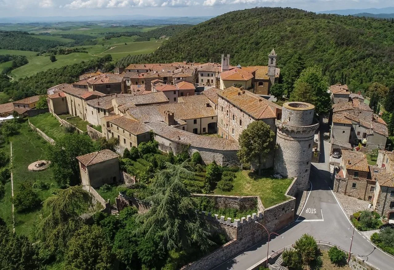 A historic village with medieval stone walls, a round tower, and red-tiled roofs, surrounded by lush green hills and farmland.