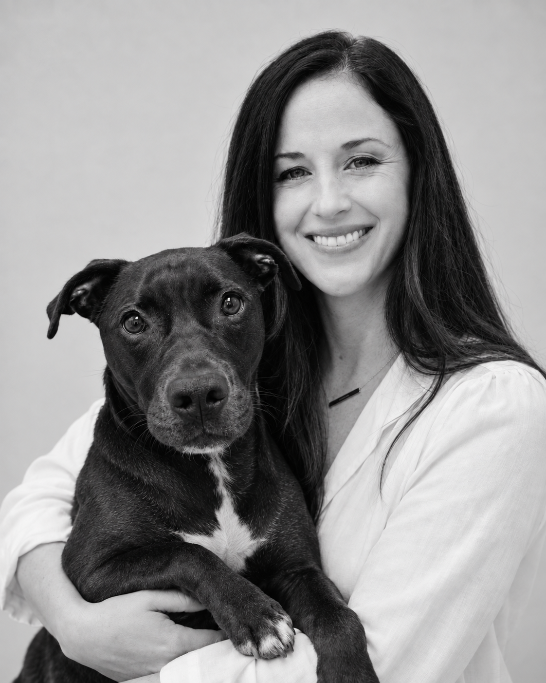 A woman with long dark hair smiling and holding a black dog with white markings on its chest. The photo is in black and white.