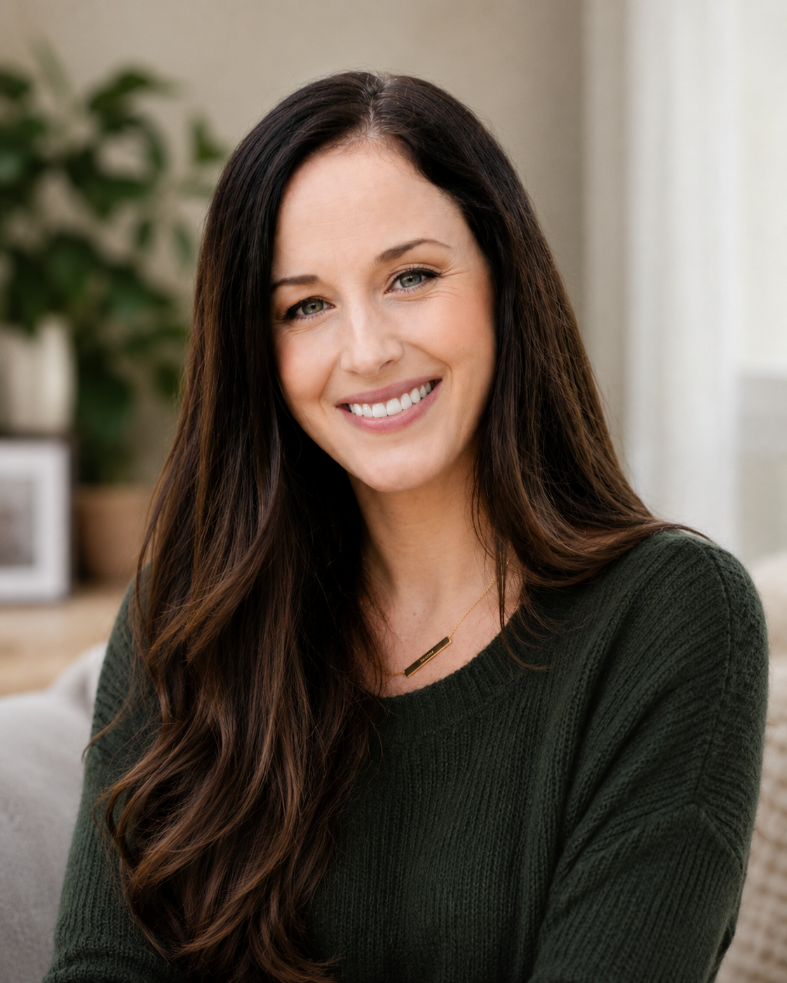 A smiling woman with long, dark brown hair and a green sweater sitting indoors near a blurred background with houseplants and framed pictures.