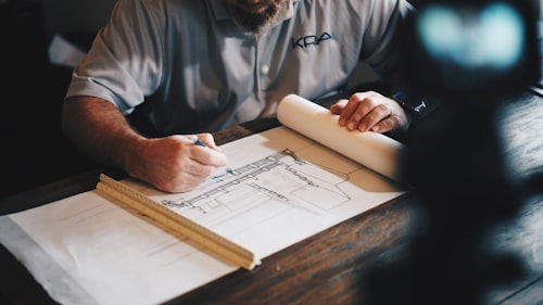 Man drawing architectural plans on blueprints with a ruler and paper on a wooden table.