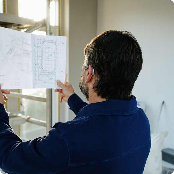 A man with dark hair holding a blueprint and pointing at it indoors near a window.