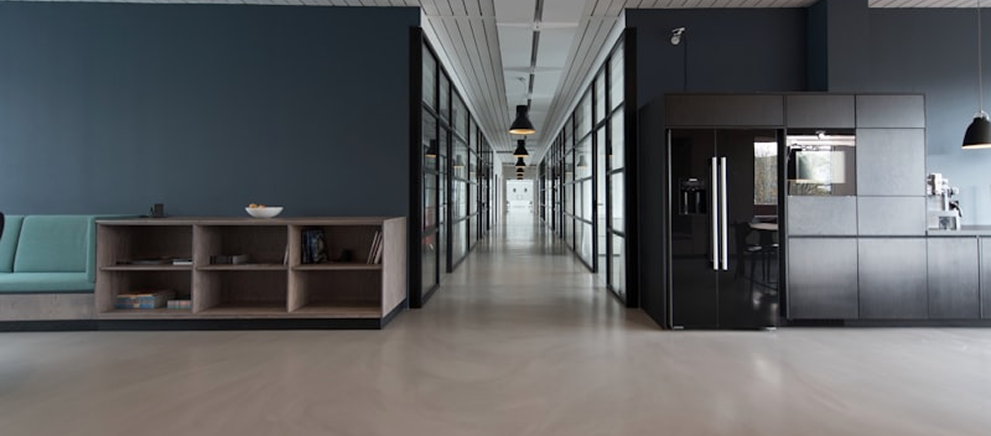 Modern office space with a hallway, glass wall rooms, a kitchen area with black cabinetry, a refrigerator, and a light-colored floor.
