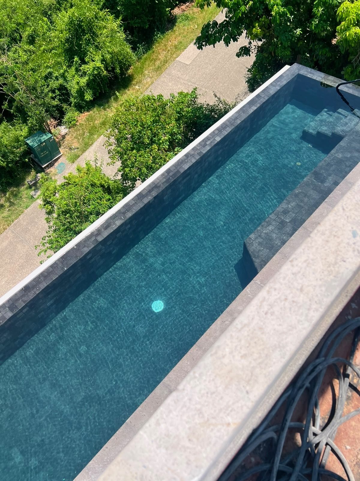 View from a balcony showing a narrow rectangular swimming pool with blue tiles and a small pool light, overlooking a backyard with green trees, bushes, a sidewalk, and a shed.