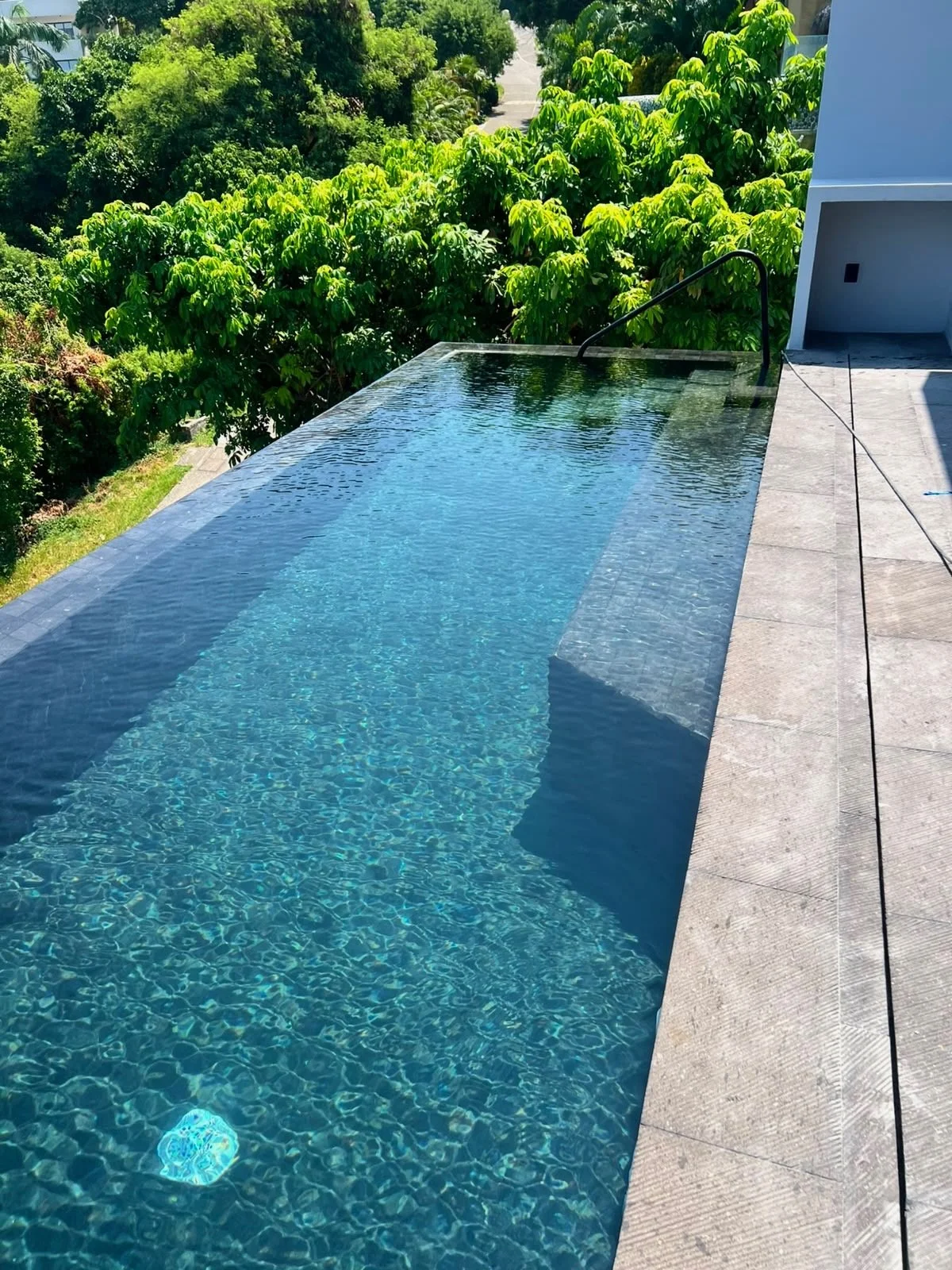 A modern infinity pool with clear blue water, adjacent to a pool deck with light gray tiles, surrounded by lush green trees and plants.