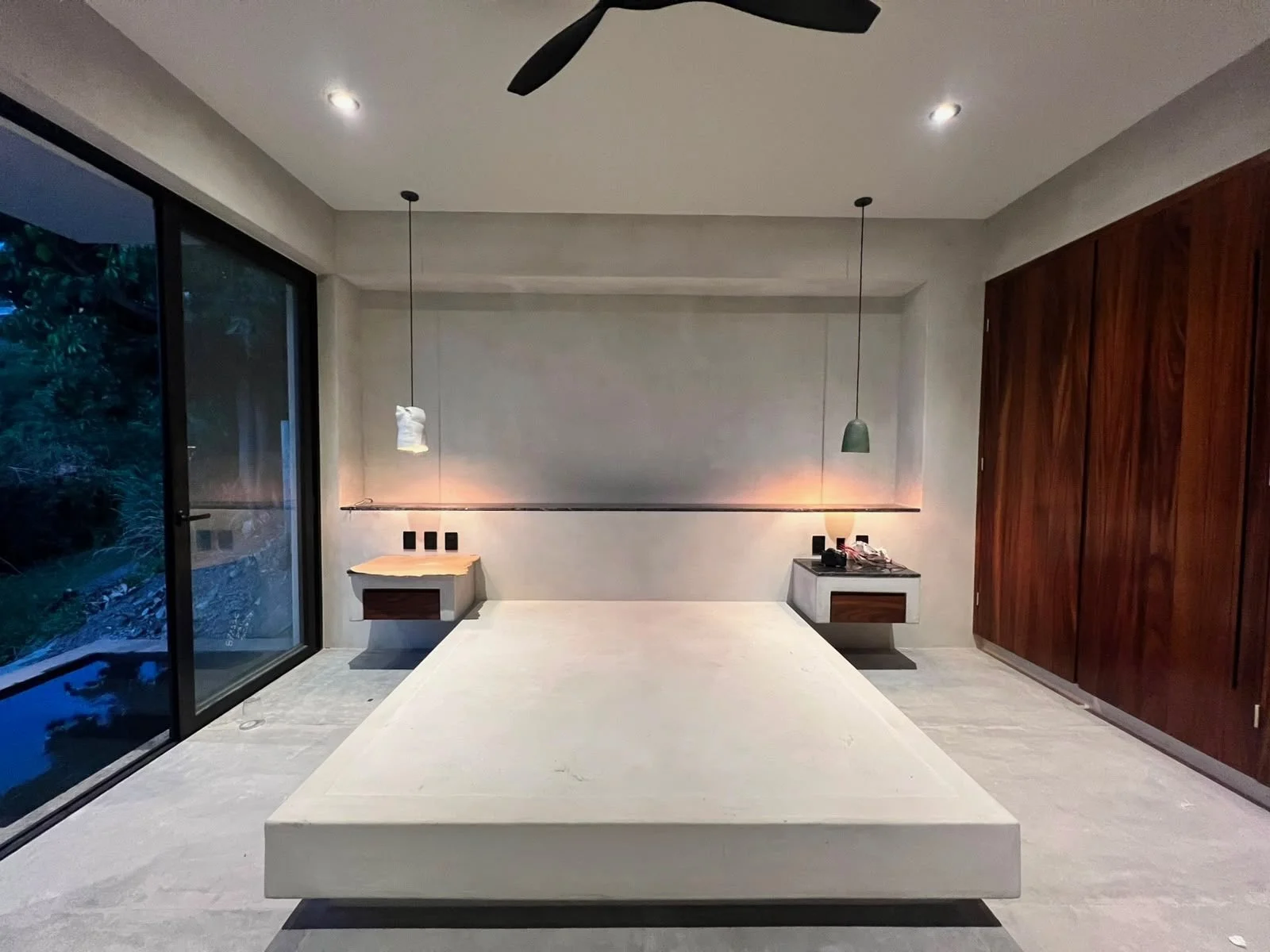 A modern minimalist bedroom with a large white bed, wood nightstands, pendant lights, a wooden closet, and a sliding glass door showing trees outside.
