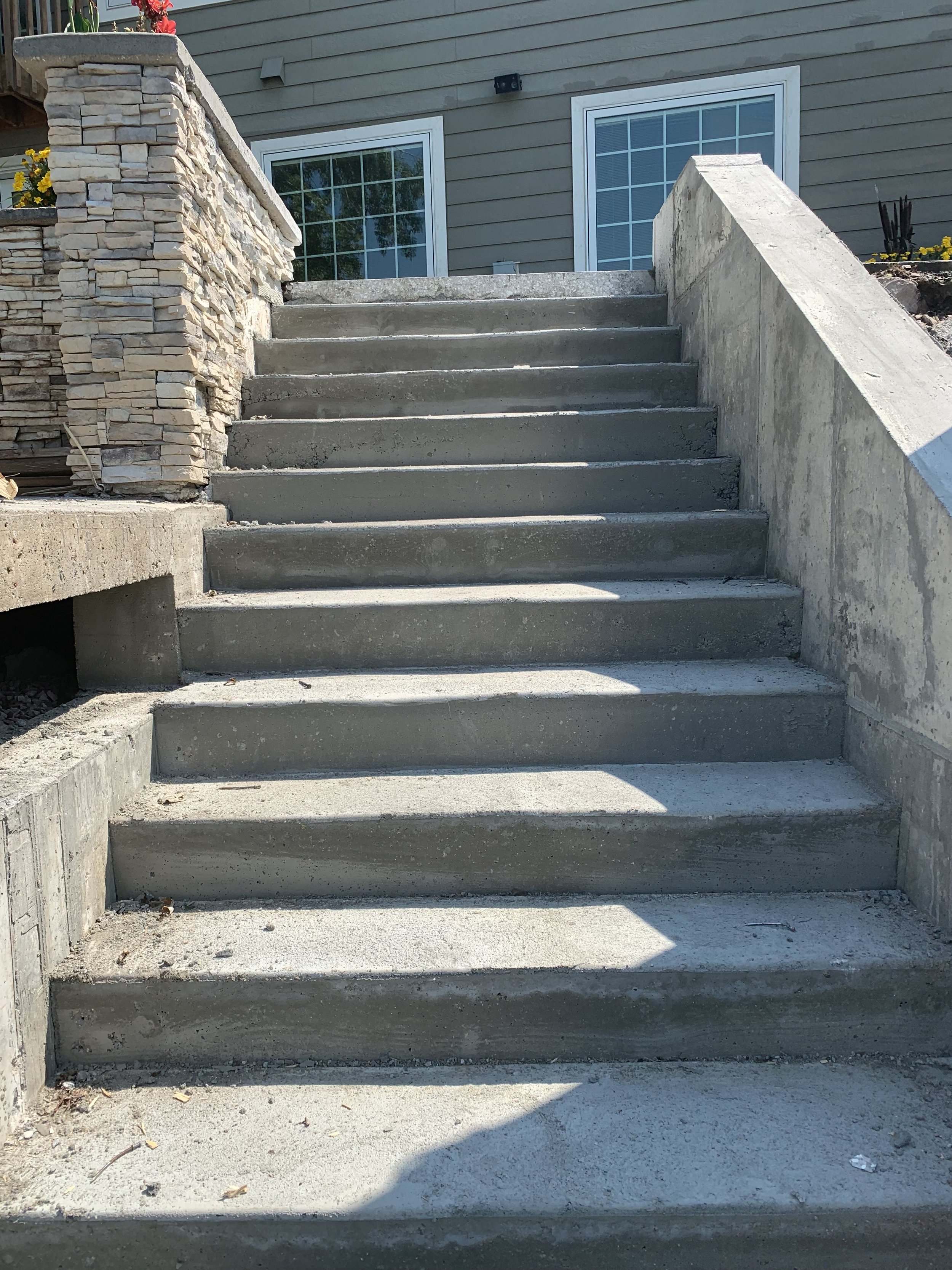 Concrete stairs leading up to a house with gray siding and large windows. There is a stone wall on the left side of the stairs and a concrete retaining wall on the right side. The stairs have visible cracks and debris around them.