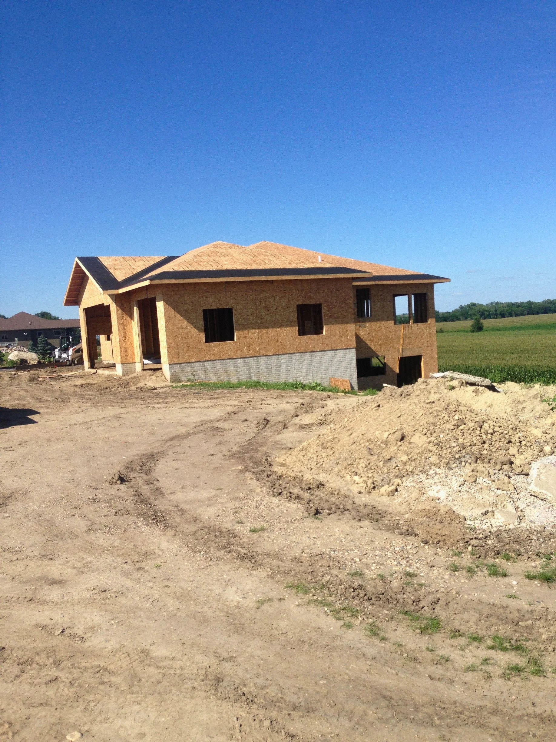 A house under construction with a partly completed exterior, missing windows and doors, set on a dirt lot with a clear blue sky overhead and green fields in the background.