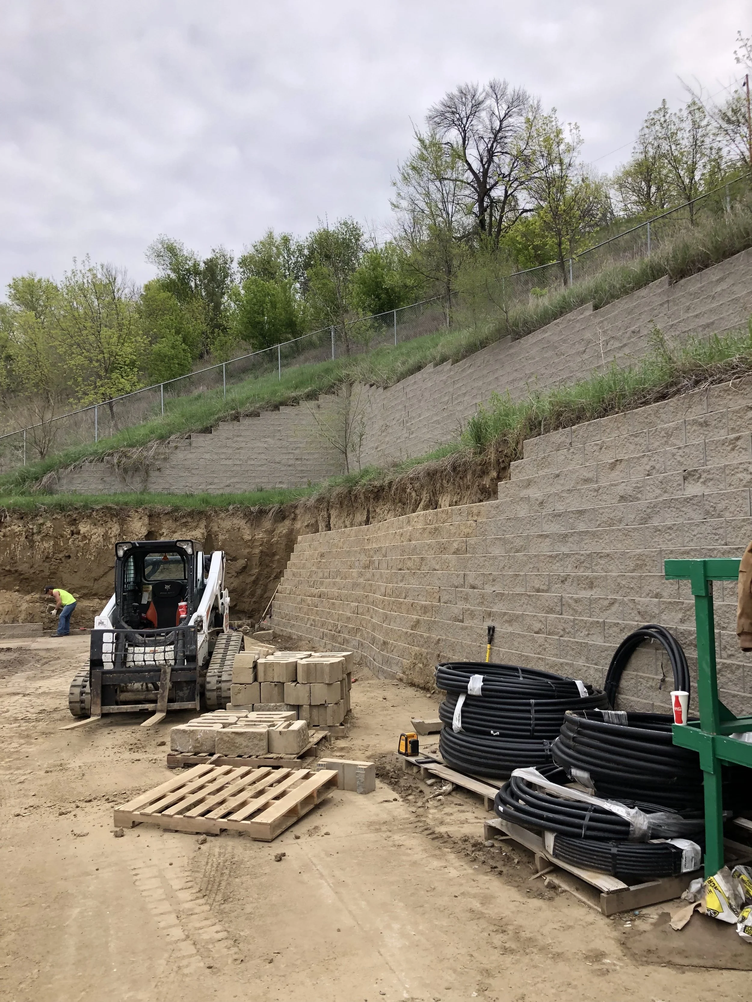 Construction site with workers and heavy machinery installing retaining walls on a hillside, with building materials and equipment scattered on dirt ground.
