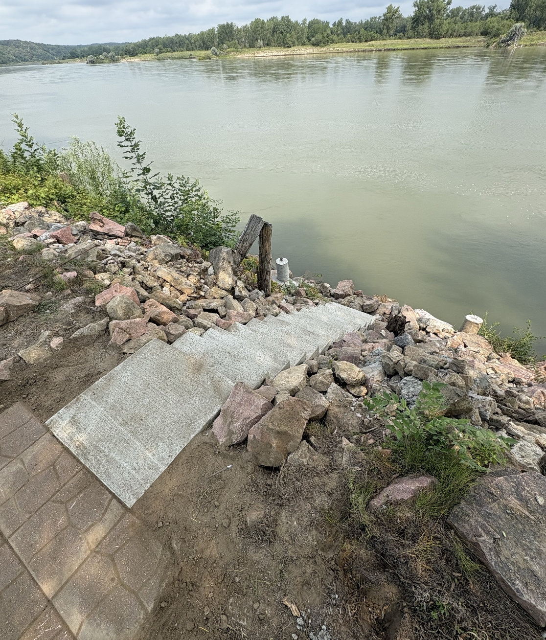 A set of concrete steps leading down to a river, with rocks and plants on either side, and a wooden post near the water. The river has a calm surface, with a tree-lined shore in the background under a cloudy sky.