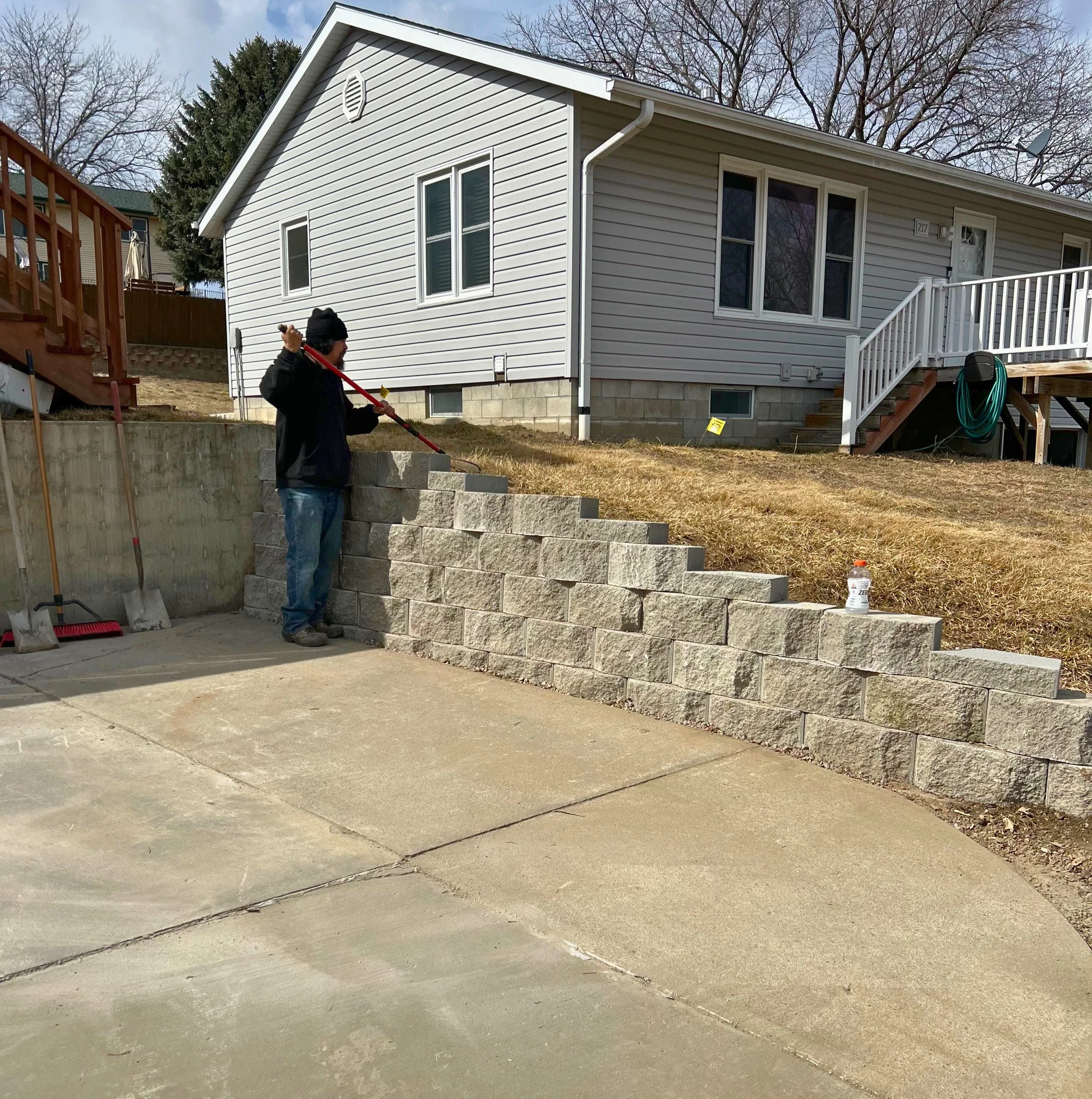 A person wearing a black jacket and hat using a shovel to move large concrete blocks in a backyard with a two-story house, stairs, and a retaining wall.