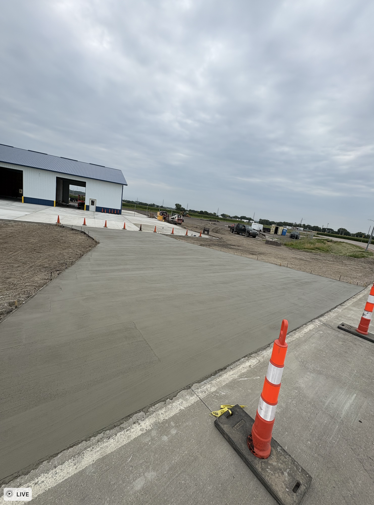 A newly poured concrete sidewalk under construction next to a building with a metal roof, with orange traffic cones and construction equipment nearby under a cloudy sky.