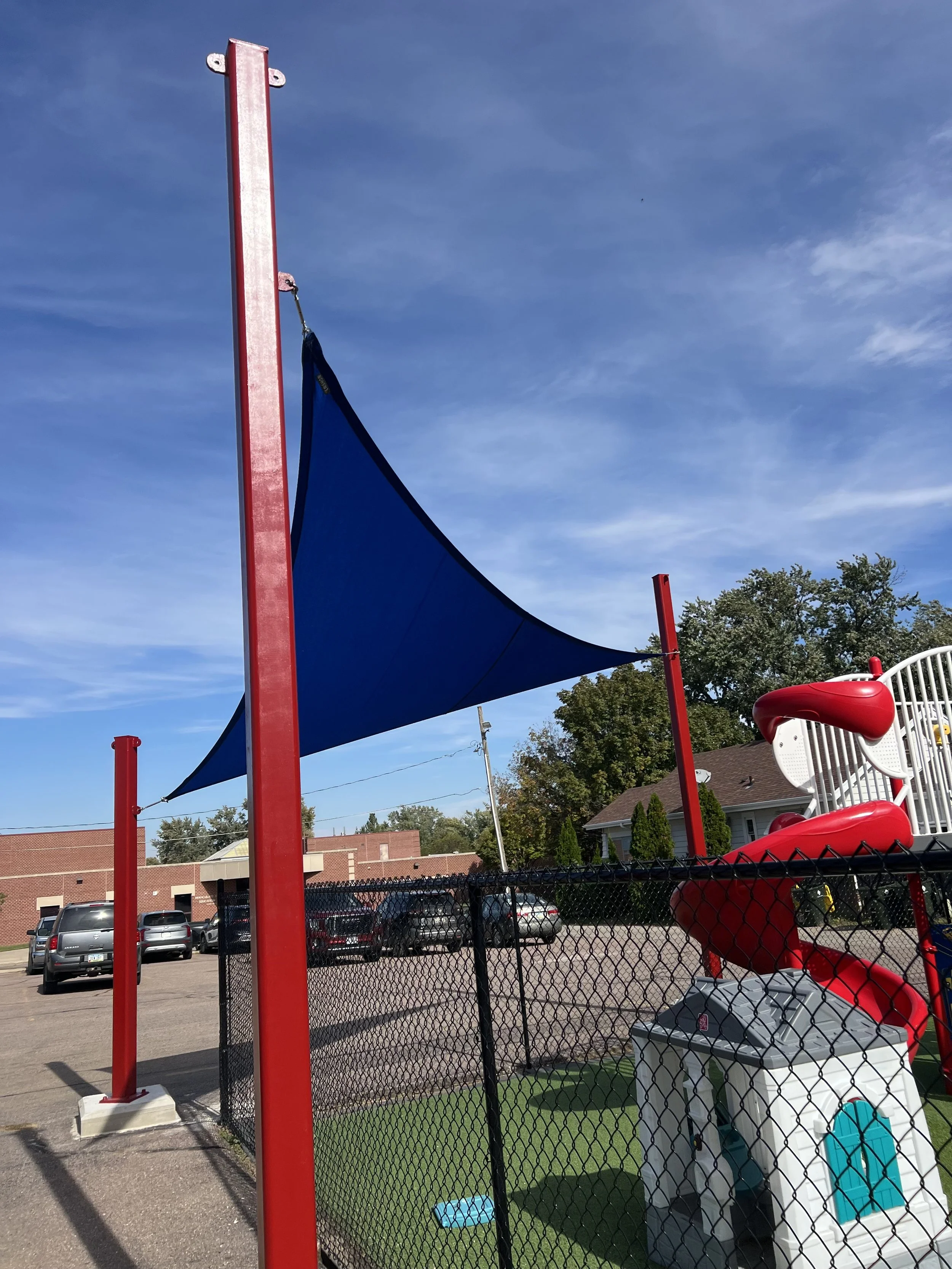 A playground area with a large blue shade sail, a red slide, and a black chain-link fence. In the background, there are parked cars, residential buildings, and trees under a blue sky with some clouds.