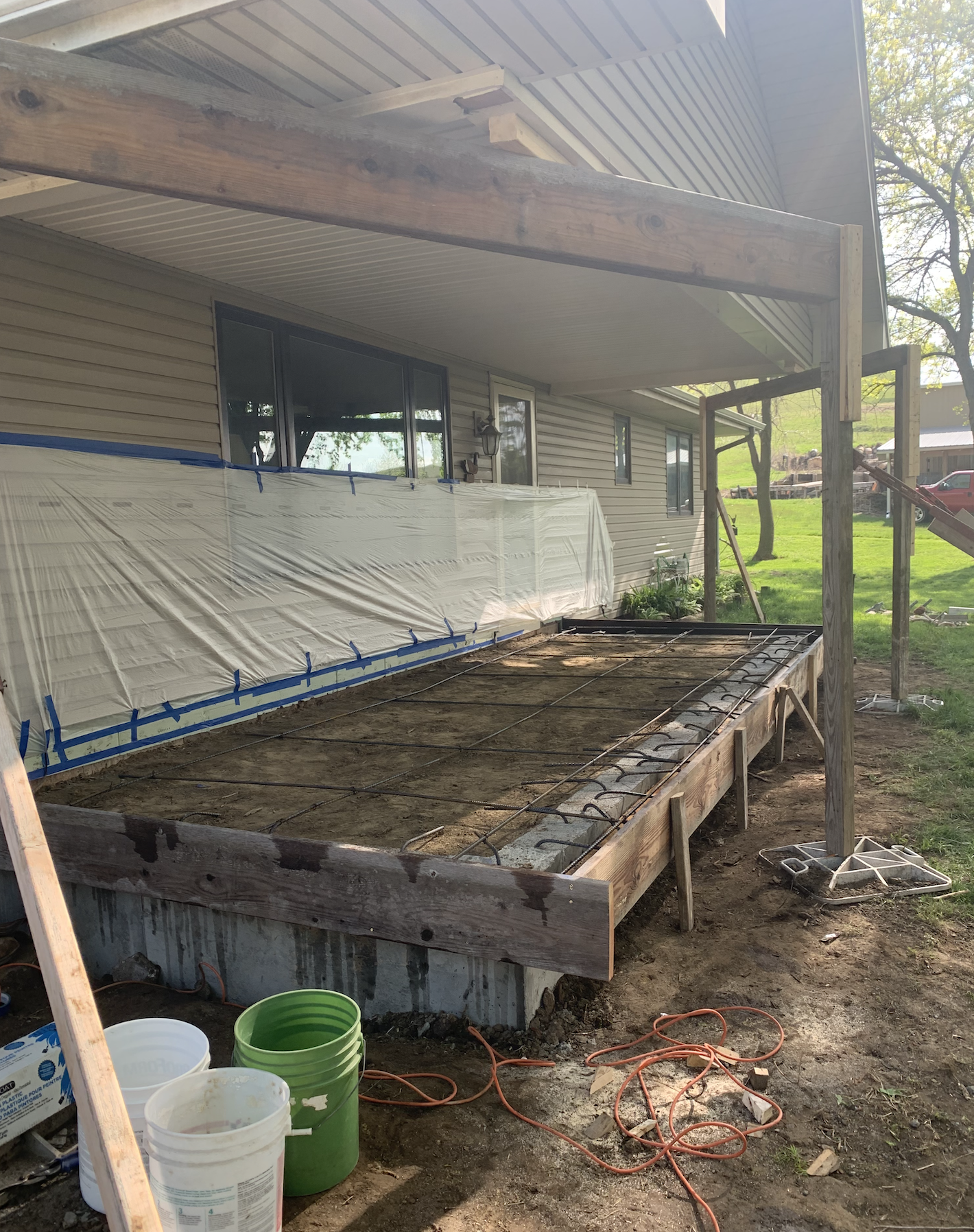 Under construction porch with wooden frame, plastic covering, and exposed ground, next to a beige house with vinyl siding and three windows, outdoors on a sunny day.