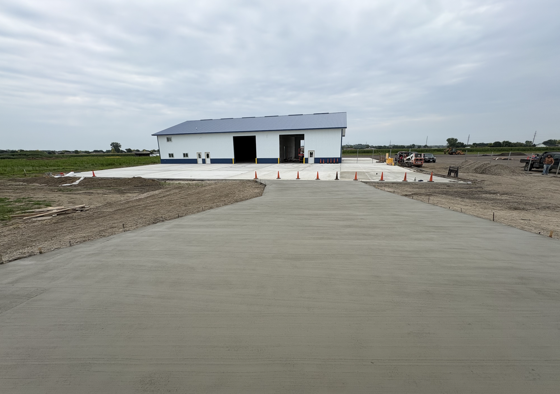 Construction site with a newly poured concrete driveway leading to a large, partially built metal building on a rural landscape under a cloudy sky.