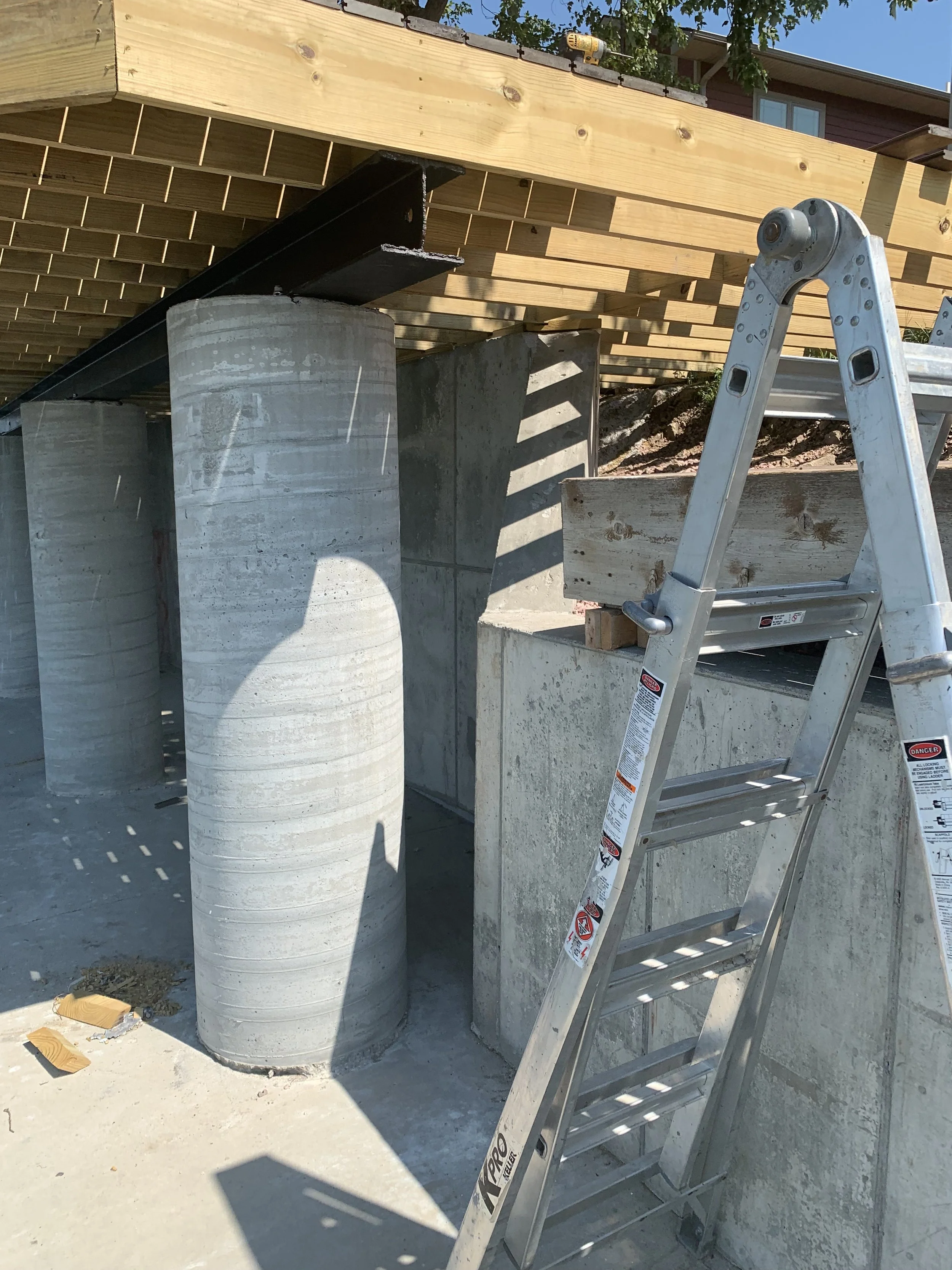 Construction site with concrete support columns, a wooden deck being built above, a aluminum ladder, and a house in the background.