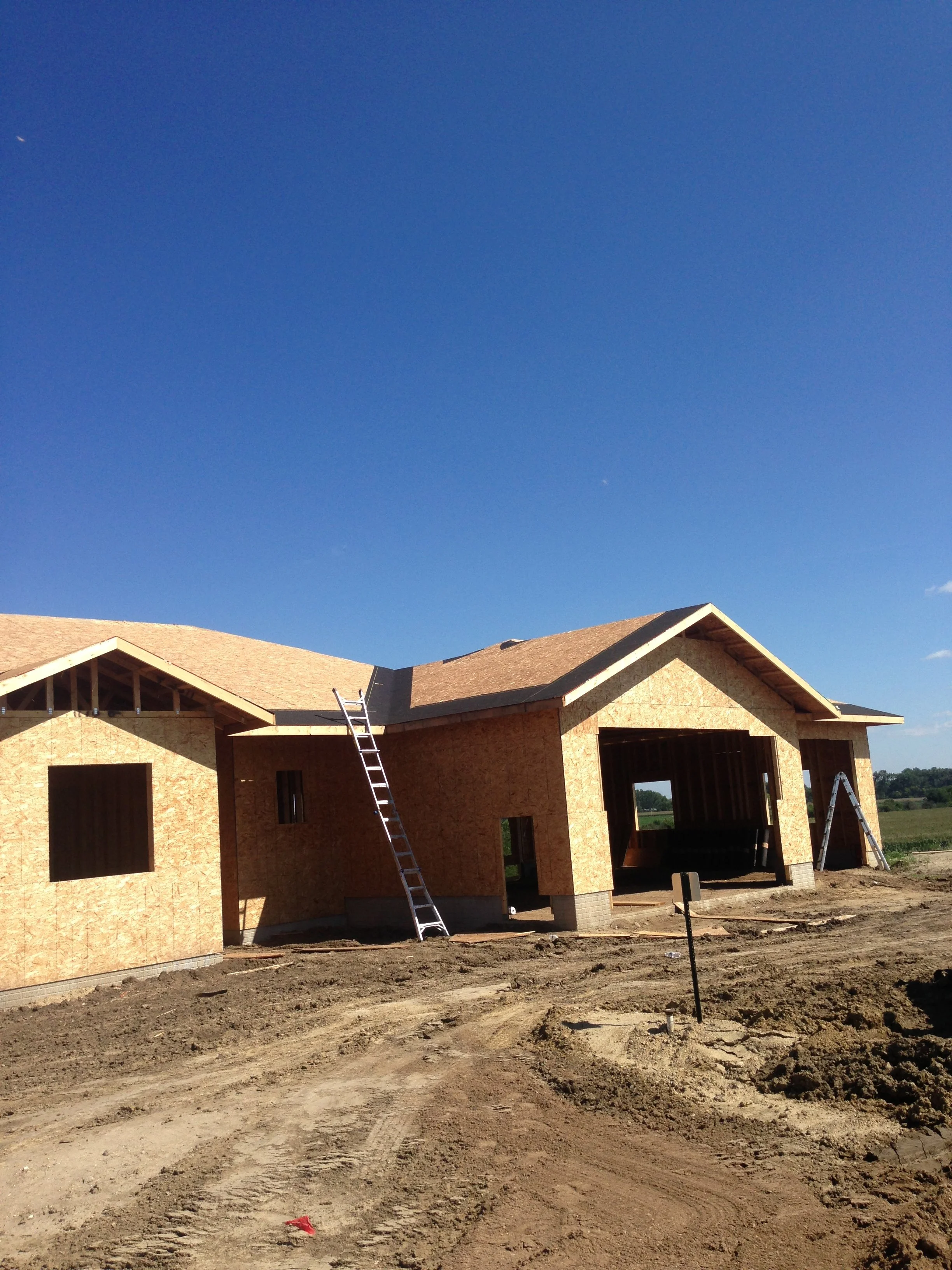 Construction site with partially built wooden houses, ladders, and dirt ground under clear blue sky.