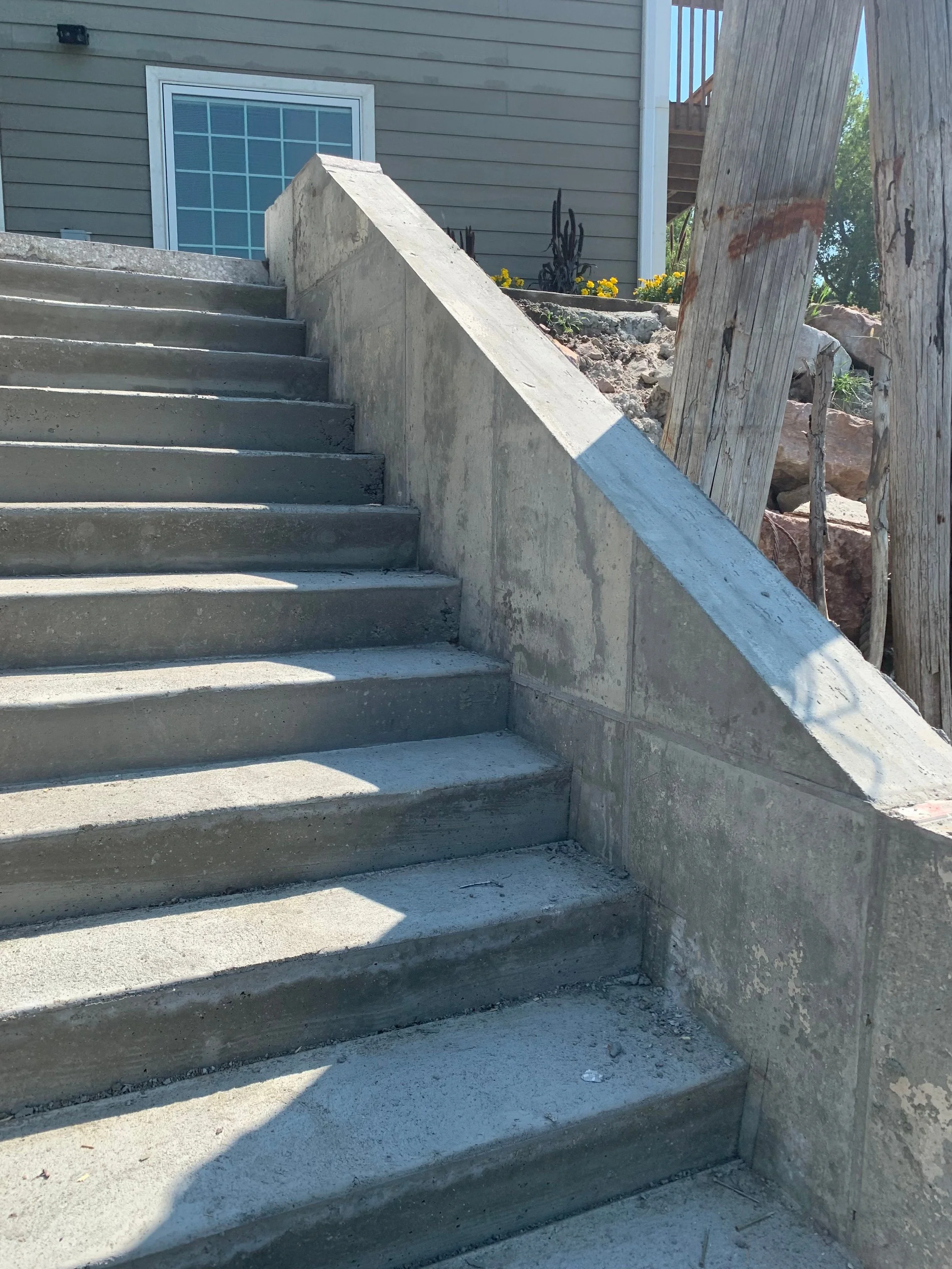 Concrete outdoor staircase with railing, leading to a house with gray siding, a window, and a backyard with garden flowers and a wooden fence.