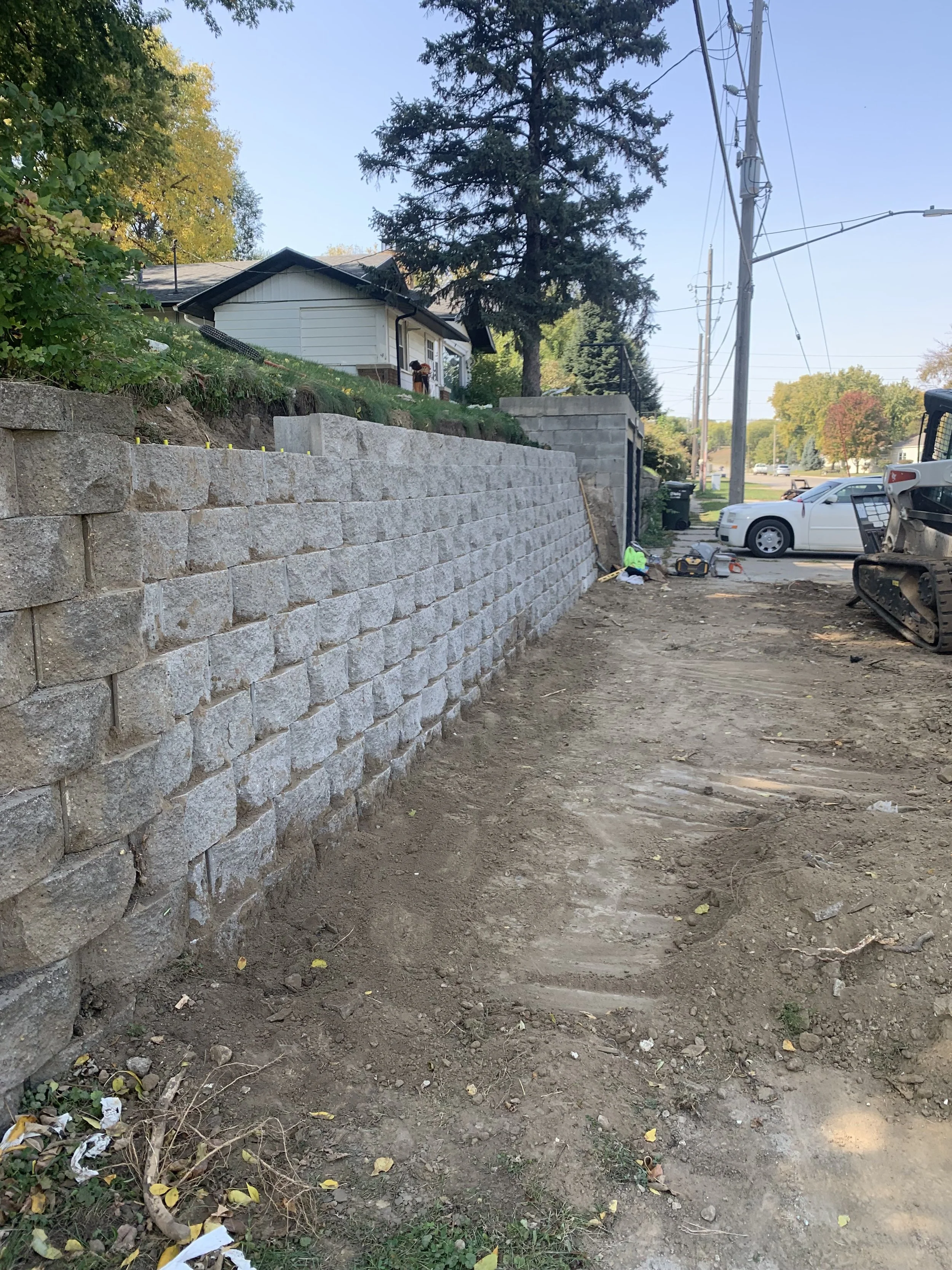 A construction site with a partially completed stone retaining wall, a dirt pathway, construction tools, and parked vehicles. Residential neighborhood with trees and power lines in the background.