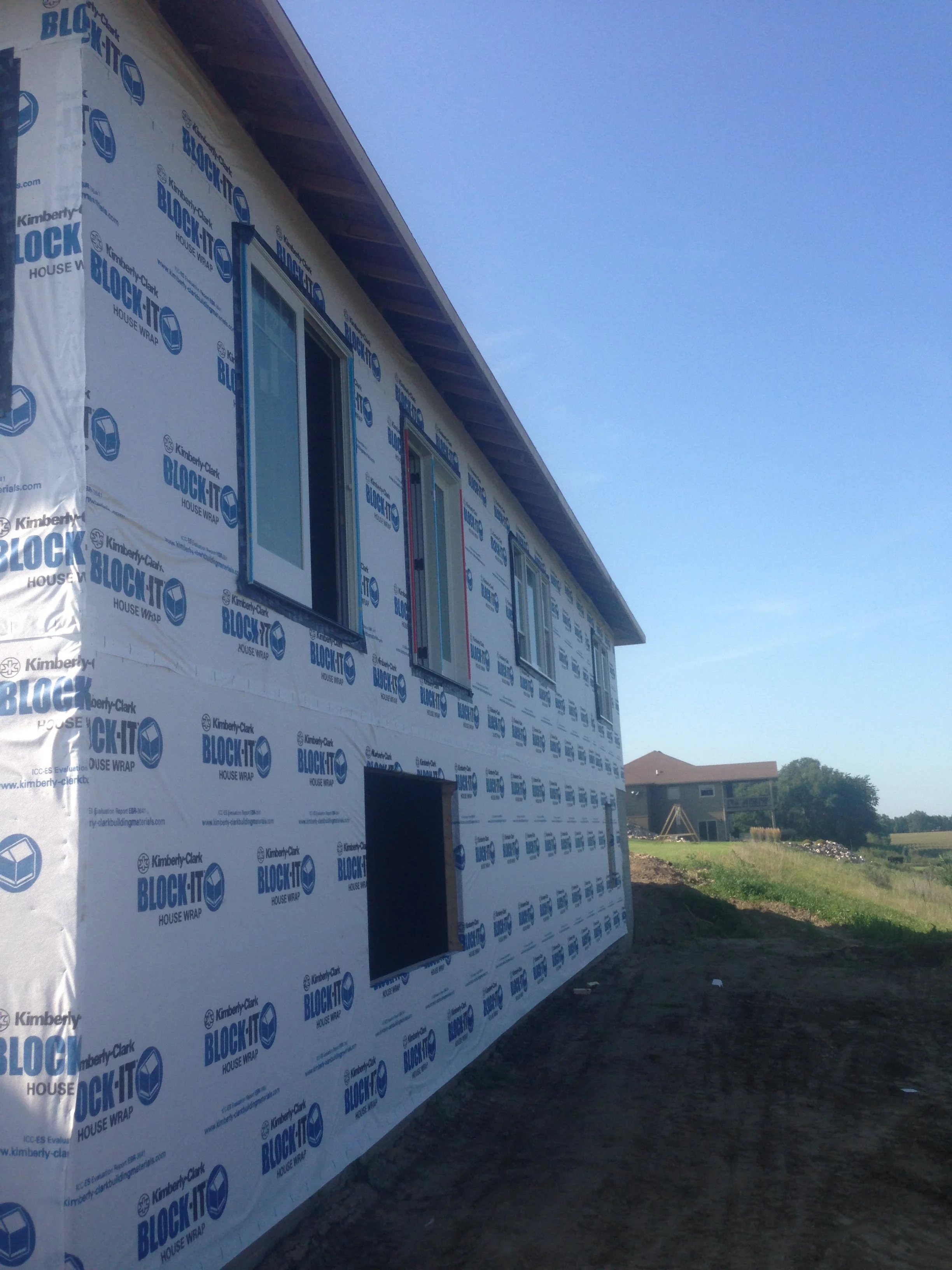 Side of a house under construction with White house wrap labeled 'BLOCK-IT' and two windows installed, at a construction site with a clear blue sky and neighboring buildings in the background.