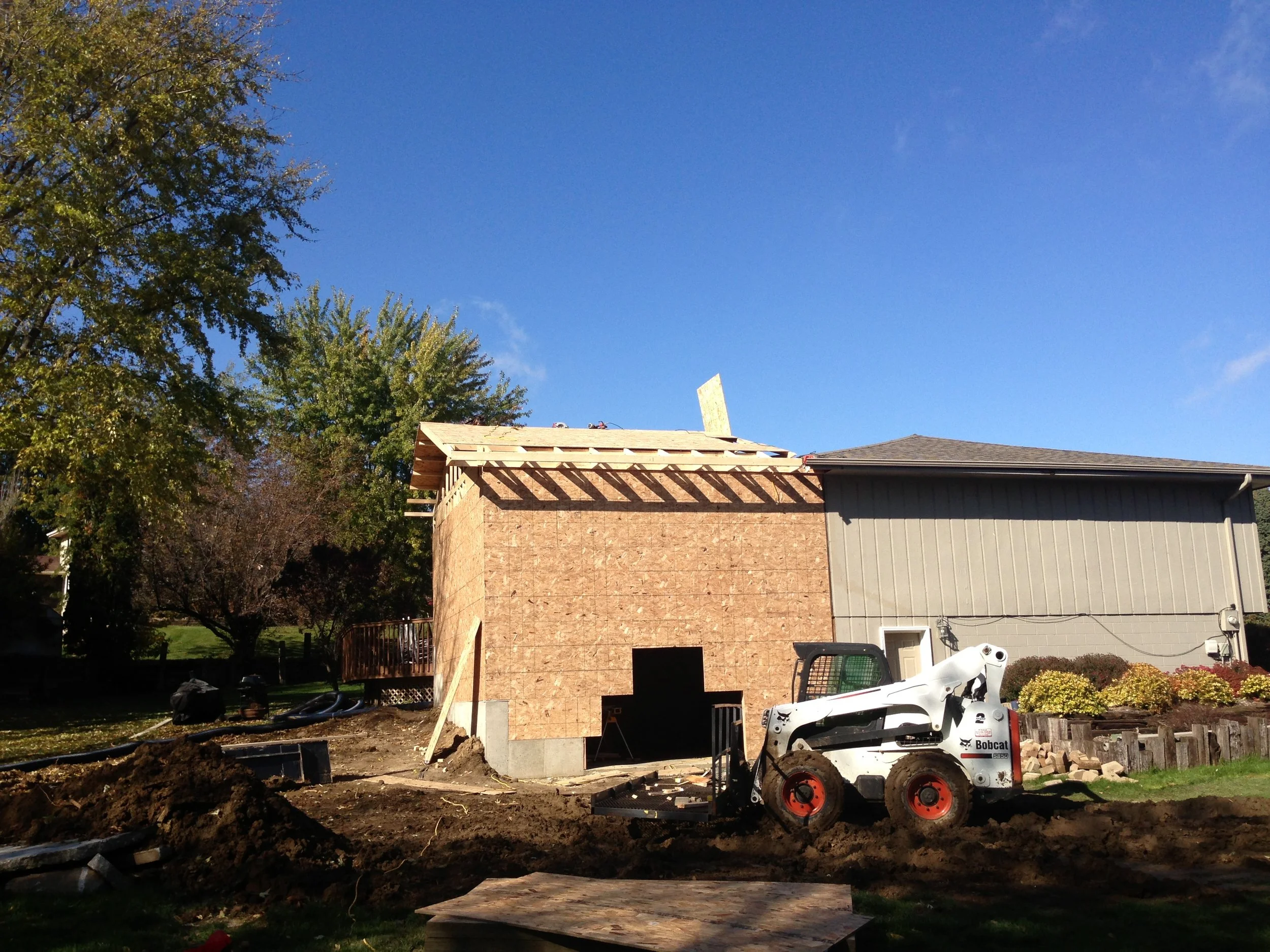 Rear view of a house under construction with wooden framing and partially installed roof, construction equipment and materials in the yard, and trees nearby under clear blue sky.