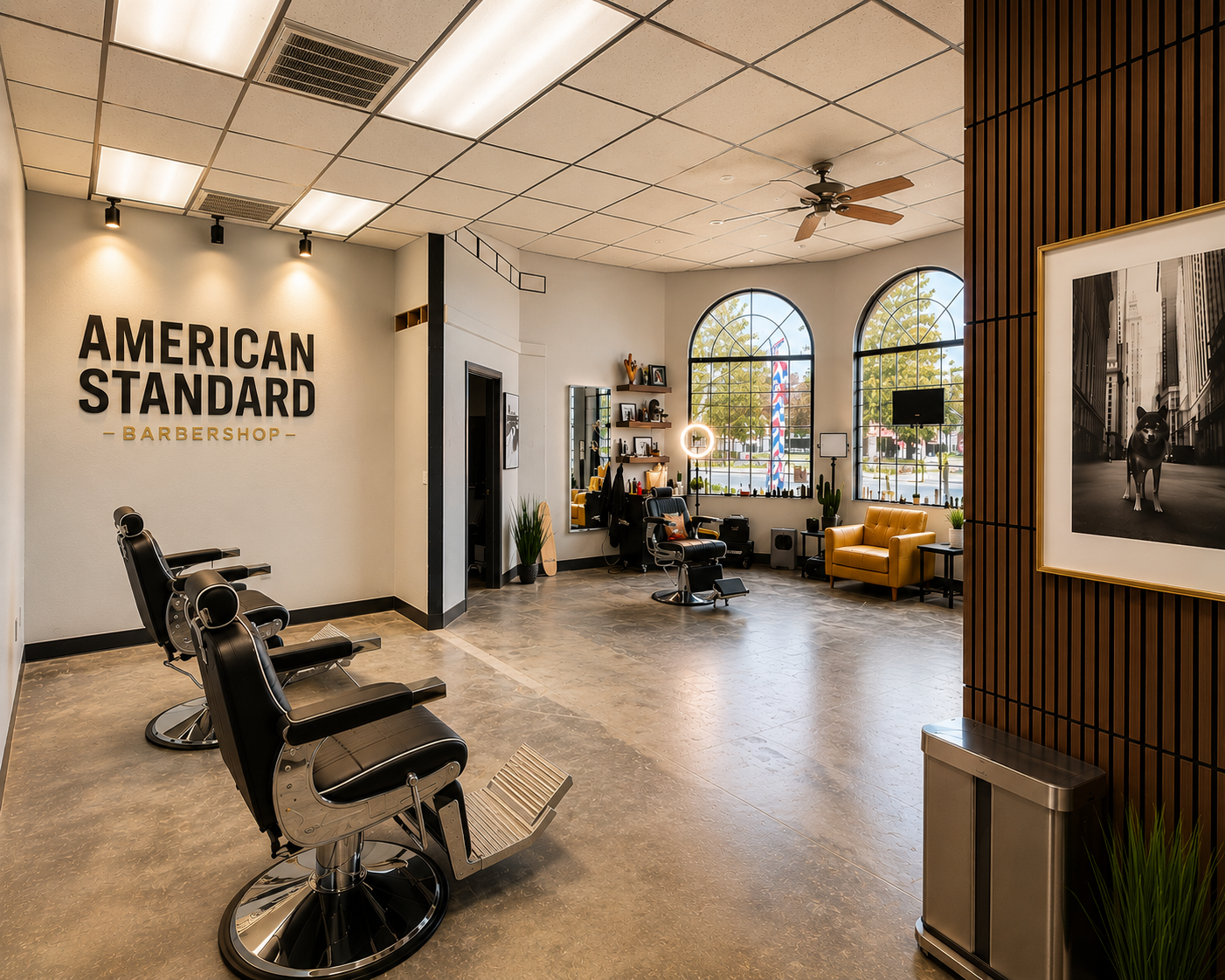 Interior of a barber shop with three black barber chairs, a yellow armchair, a large framed photograph of a dog on a city street, and a waiting area with shelves and decorative plants near large windows.