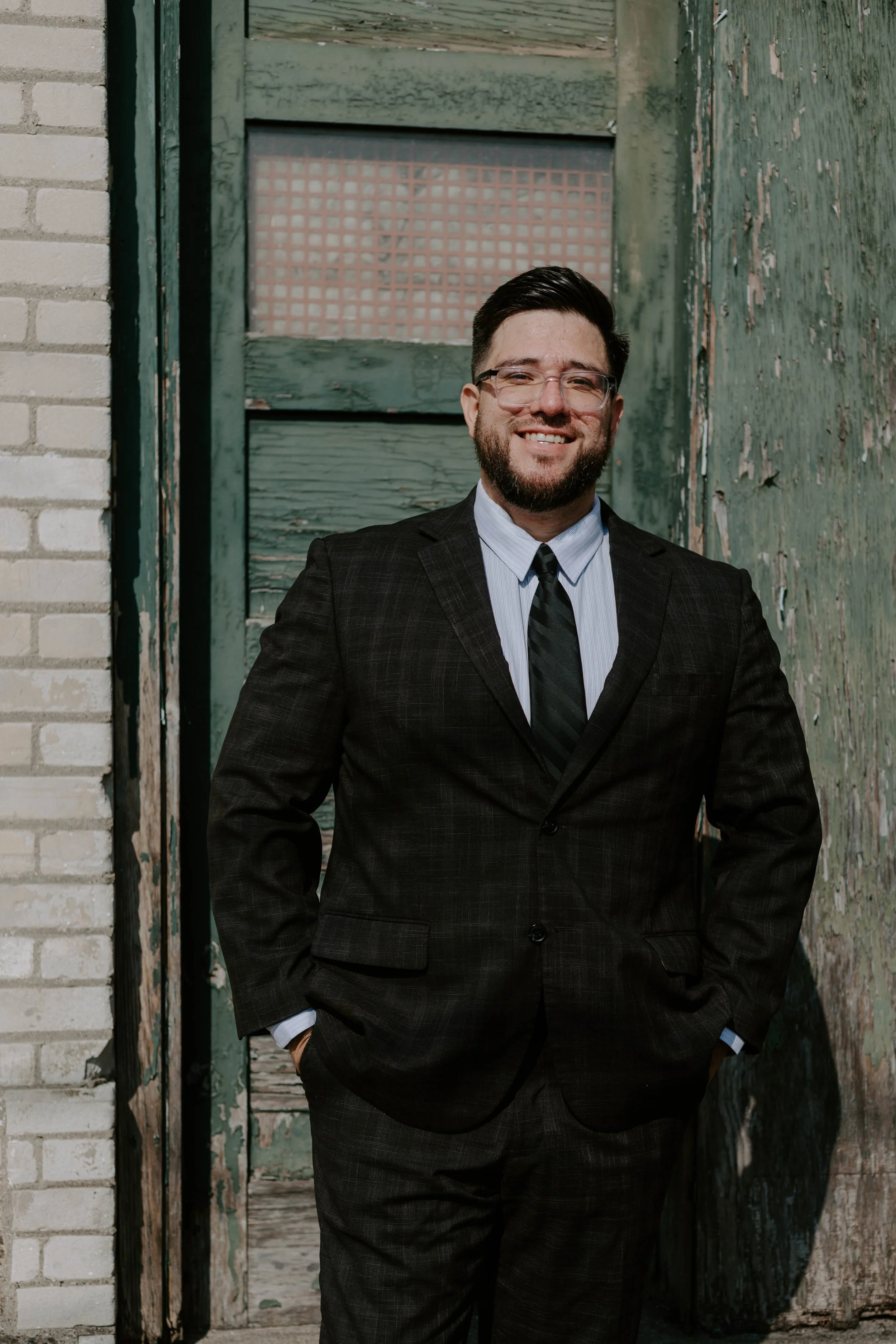 A man in a dark suit and striped tie, wearing glasses, standing in front of a weathered green wooden door with peeling paint, and a brick building wall on the left.