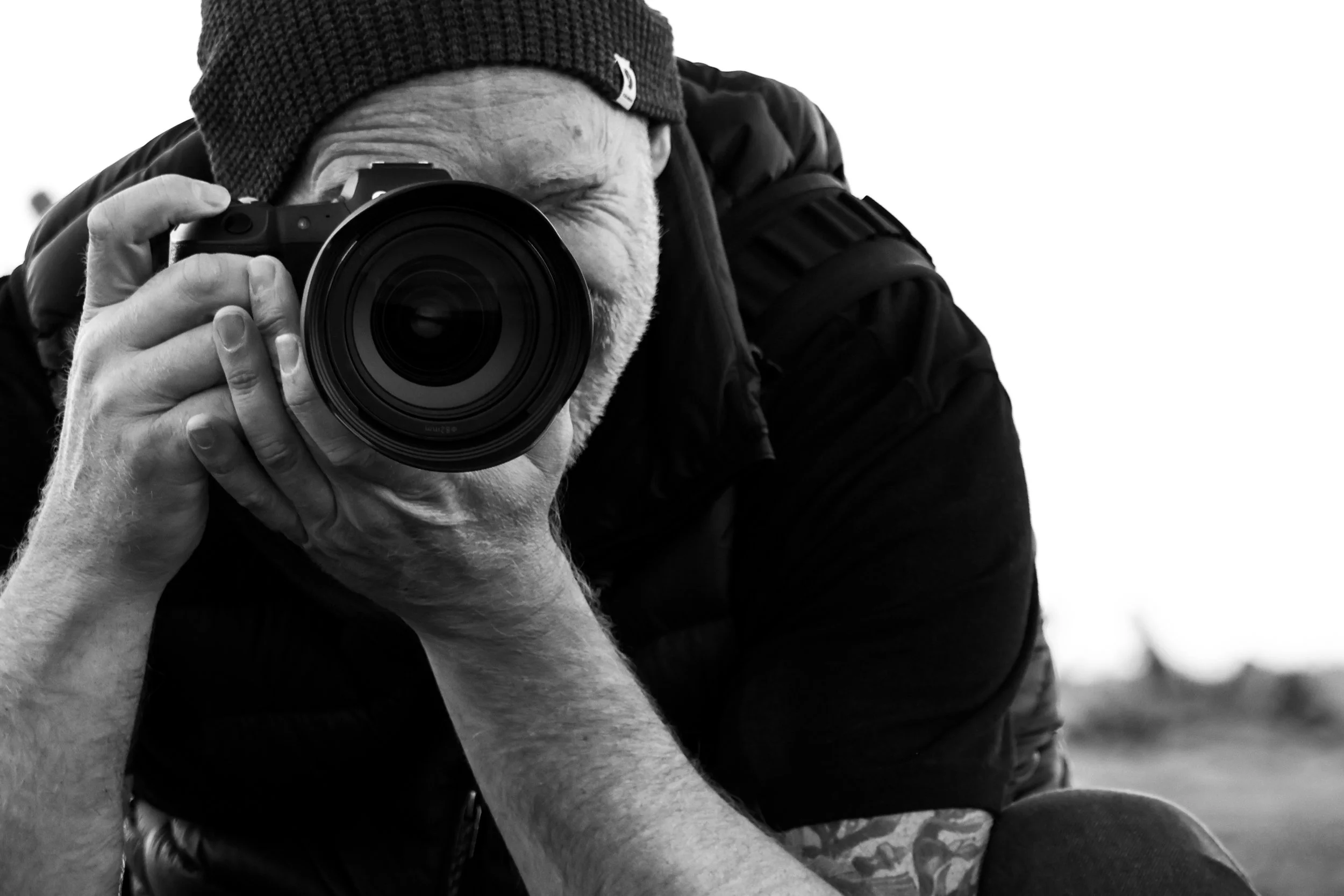 Photographer Cameron Griffin wearing a knit cap taking a black and white photo with a camera, focusing through the camera's lens.