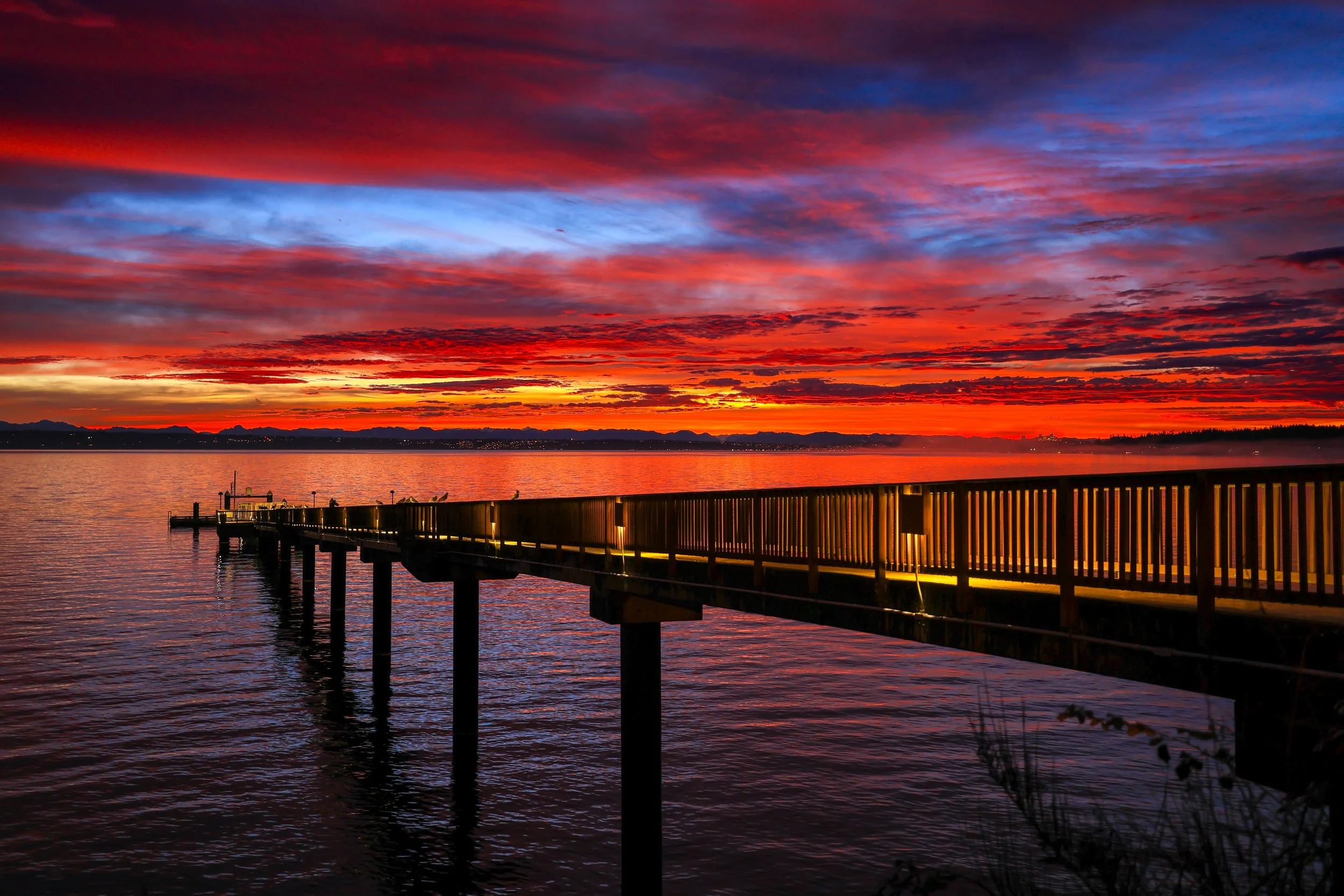 Sunrise over Puget Sound with vibrant red, orange, pink, and blue hues in the sky, and the Suquamish wooden pier extending into the water near Bainbridge Island