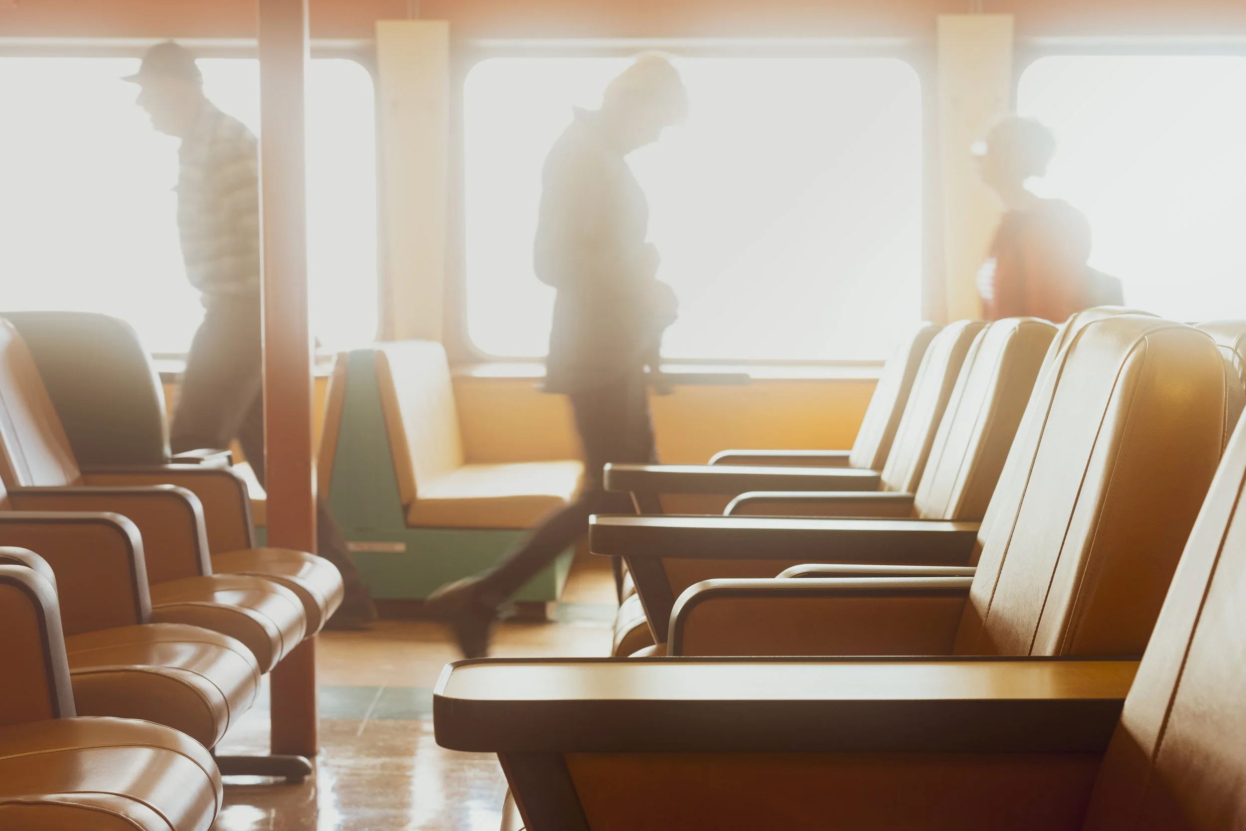 Silhouettes of three people walking through a Bainbridge-Seattle ferry backlit by bright sunlight from large windows.