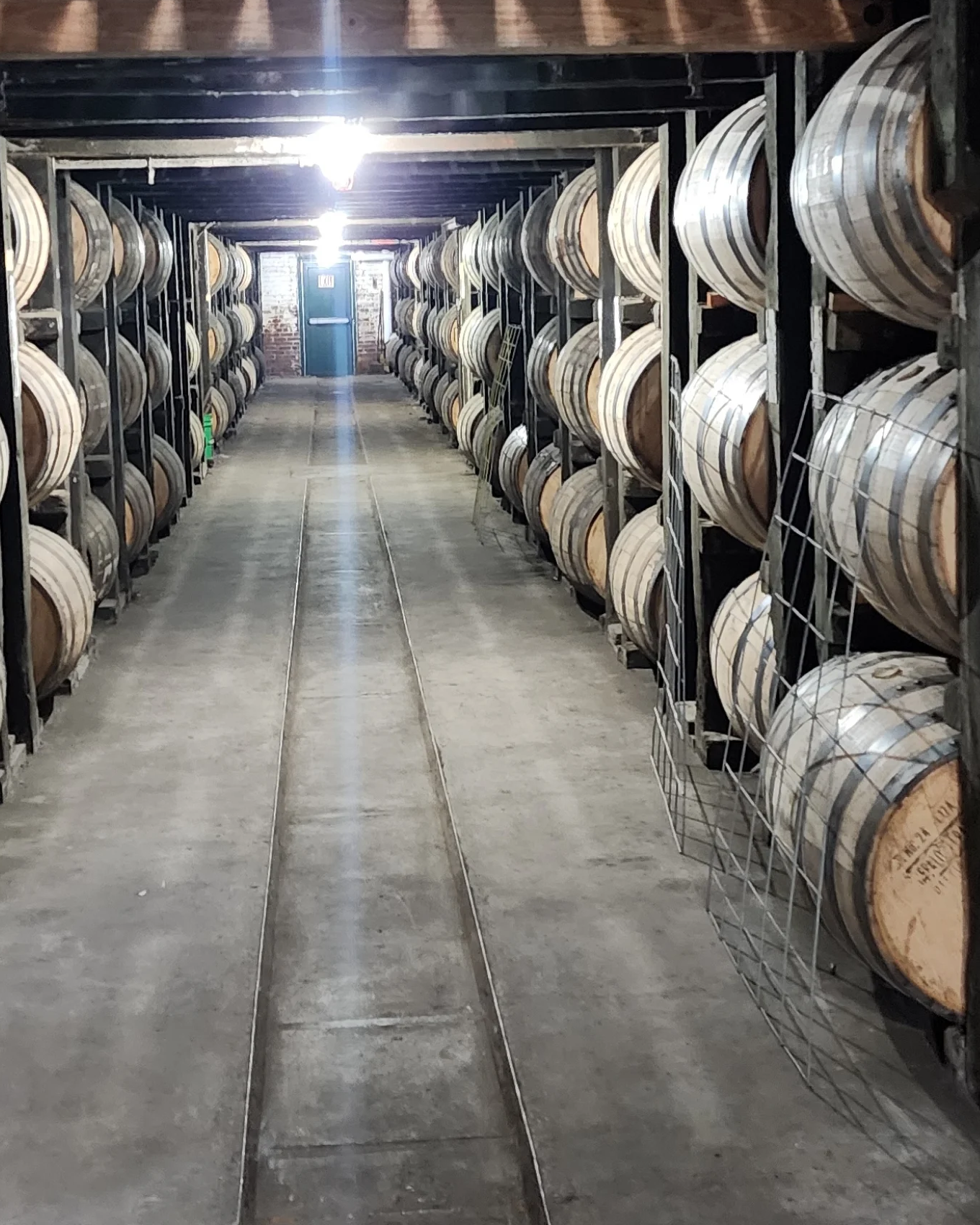 Rows of wooden barrels stored on metal racks in a cellar with a concrete floor, brick walls, and a blue door at the end, illuminated by bright ceiling lights.
