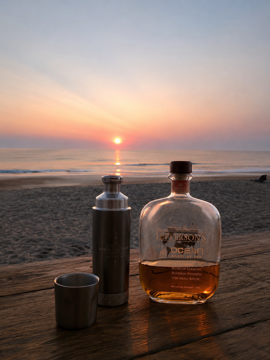 A bottle of Jefferson's Ocean whiskey, a small metal cup, and a thermos on a wooden surface at the beach during sunset, with the ocean and sunset in the background.