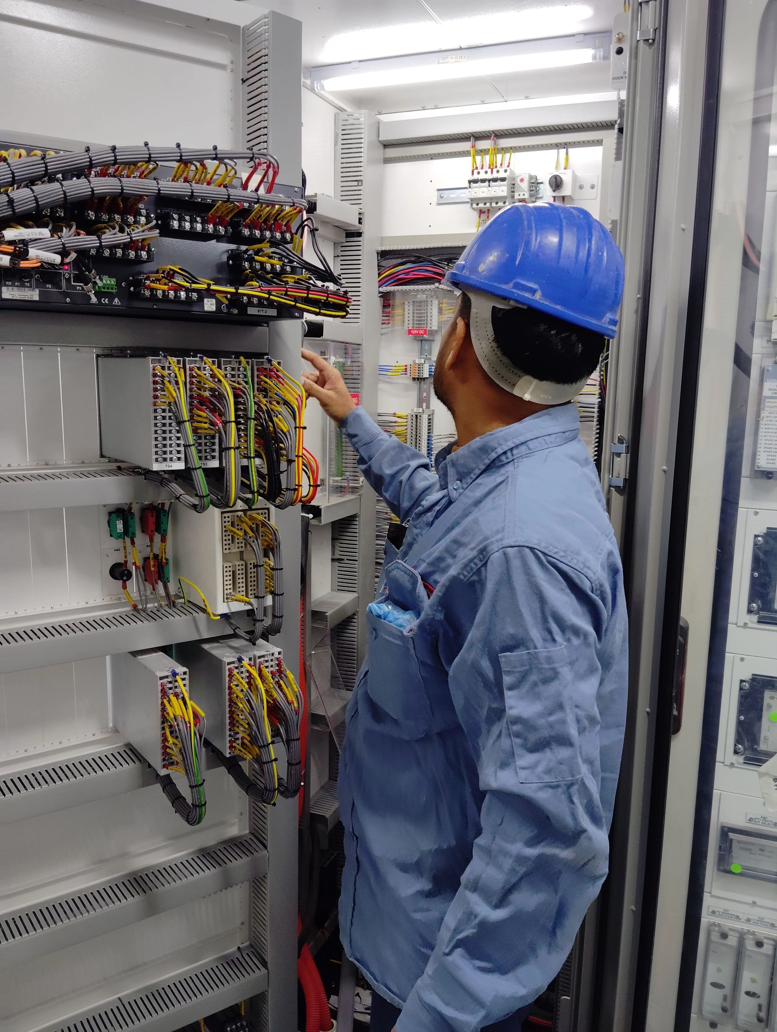 A technician wearing a blue safety helmet and a denim shirt inspecting electrical wiring and equipment inside a control panel or electrical cabinet.