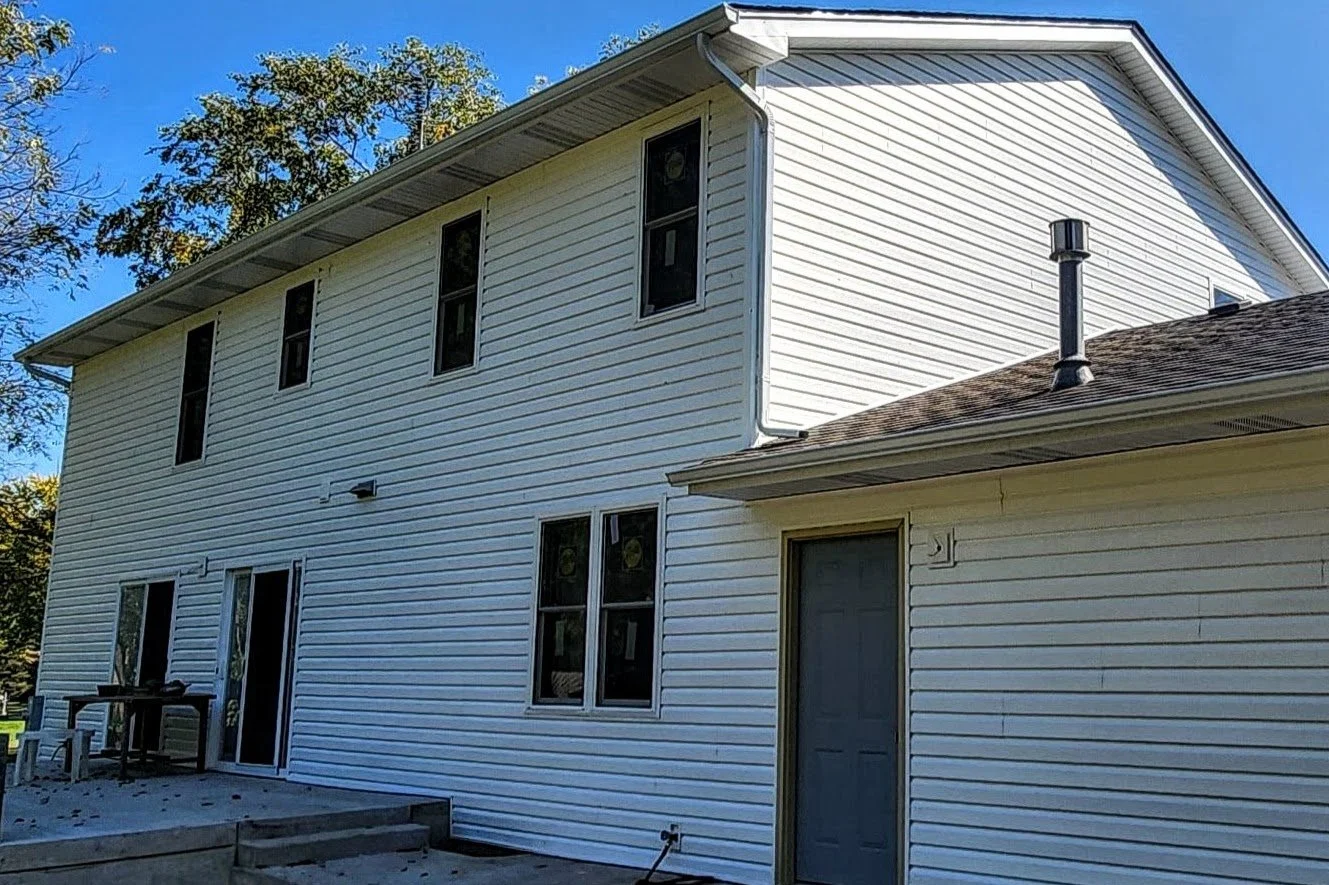 A two-story house with white vinyl siding, multiple windows, and a small patio area with a table and chairs outdoors. Trees and a clear blue sky are visible in the background.