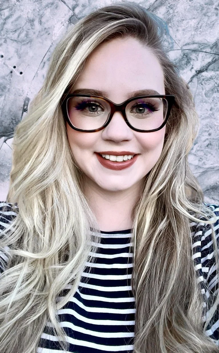 A young woman with long blonde hair, wearing glasses and a black and white striped shirt, smiling at the camera with a gray marble background.