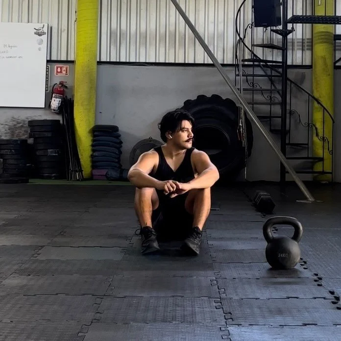 Young man sitting on the gym floor in workout clothes next to a kettlebell, in a gym with stacked weights, tires, and gym equipment in the background.