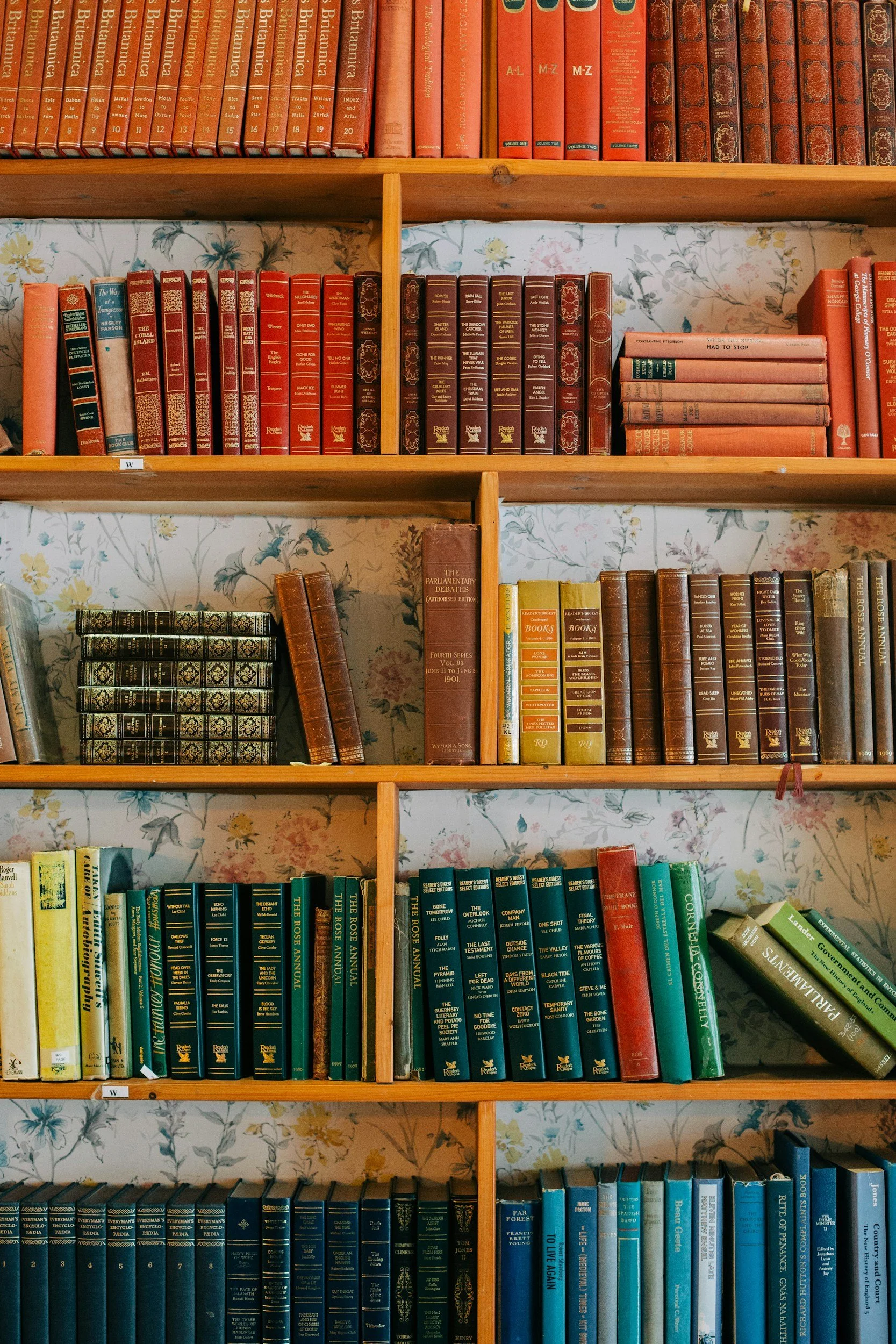 Wooden bookshelf filled with various colored books, some stacked horizontally and others vertically, against a floral wallpaper background.