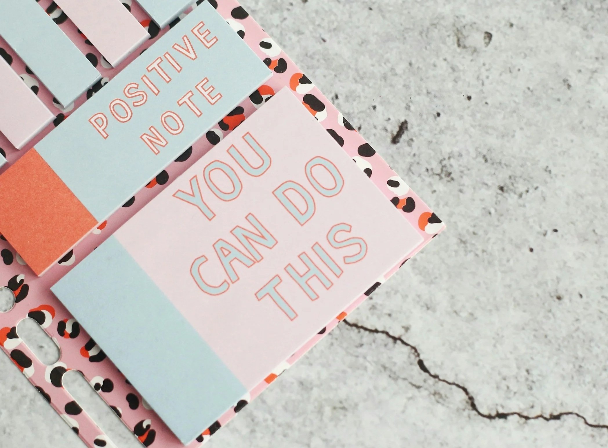Close-up of colorful greeting cards on a cracked gray stone surface. The cards are pink and light blue with playful print and text.