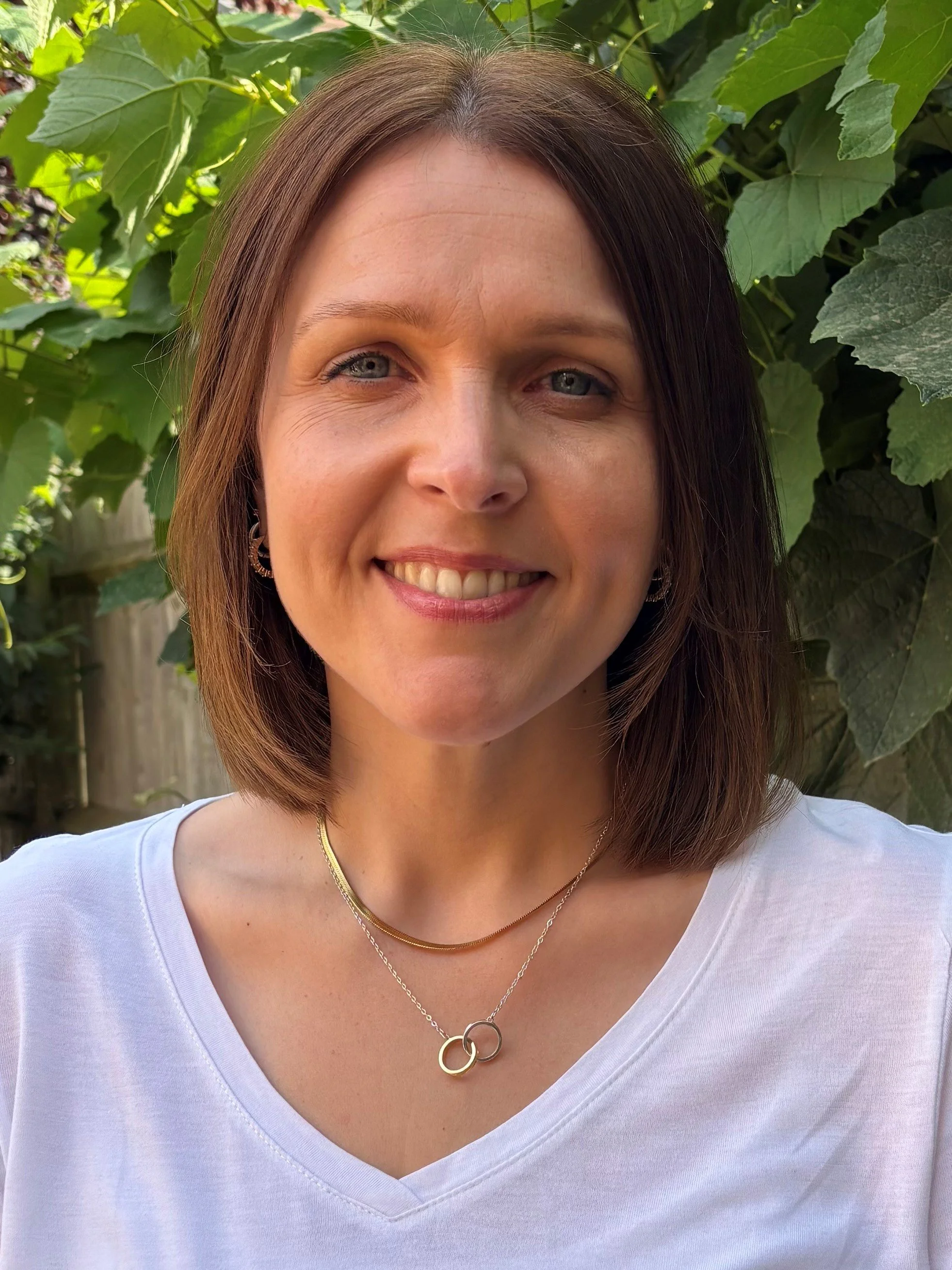 Close-up of a woman with shoulder-length brown hair, smiling, wearing a white V-neck shirt, gold necklaces, and earrings, standing outdoors in front of green leafy plants.