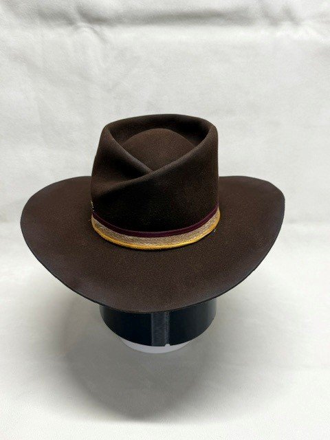 Brown cowboy hat with a decorative band, displayed on a black stand against a white background.