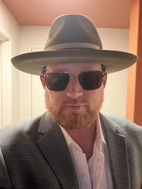A man with a beard wearing a gray hat, sunglasses, a gray blazer, and a white shirt.