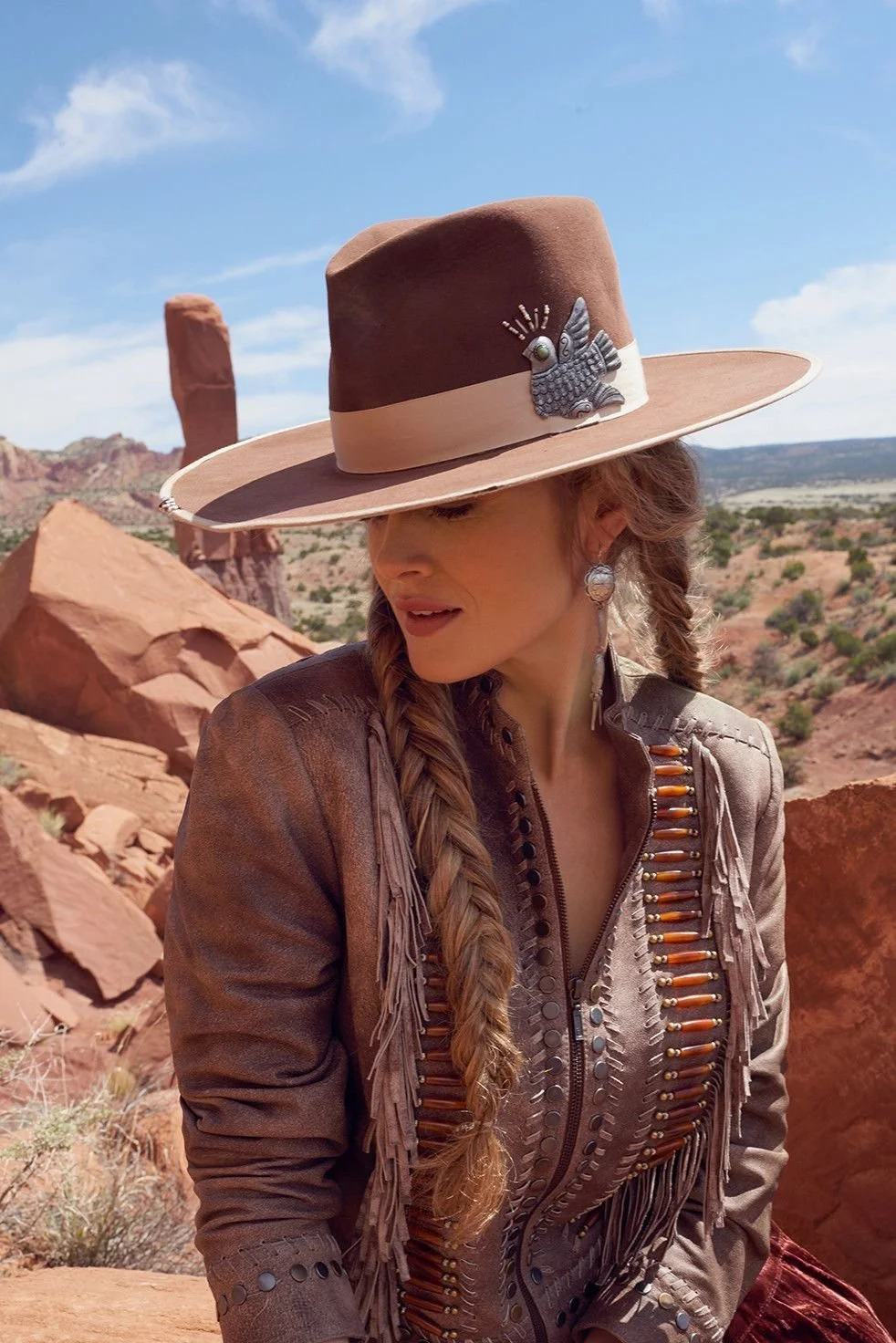 Woman wearing a tan cowboy hat with a feather and bird embellishment, dressed in a fringed leather jacket with metal details, sitting in a rocky desert landscape with red rock formations.