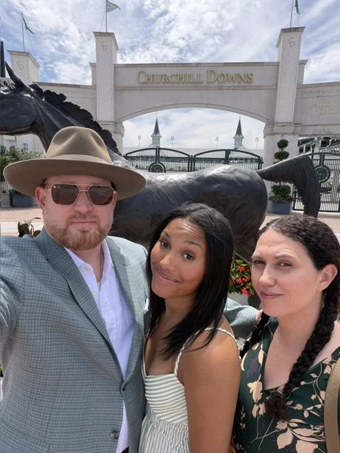Three people taking a selfie in front of Churchill Downs with the entrance arch and a horse statue in the background.