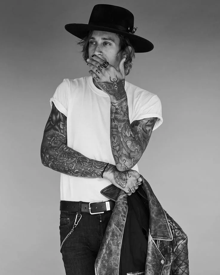 Black and white photo of a young man with tattoos, wearing a white t-shirt, a wide-brimmed hat, and holding a jacket. He has his hand covering his mouth and appears contemplative.